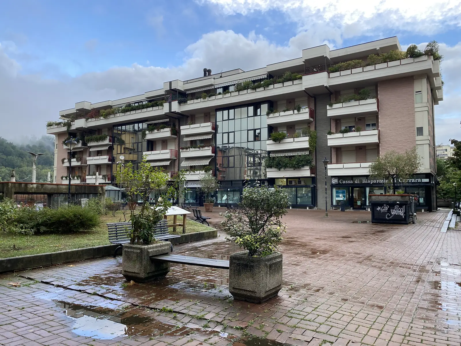 A wet paved square with benches and potted shrubs faces a balconied apartment block housing shops, including a branch of Cassa di Risparmio di Carrara.
