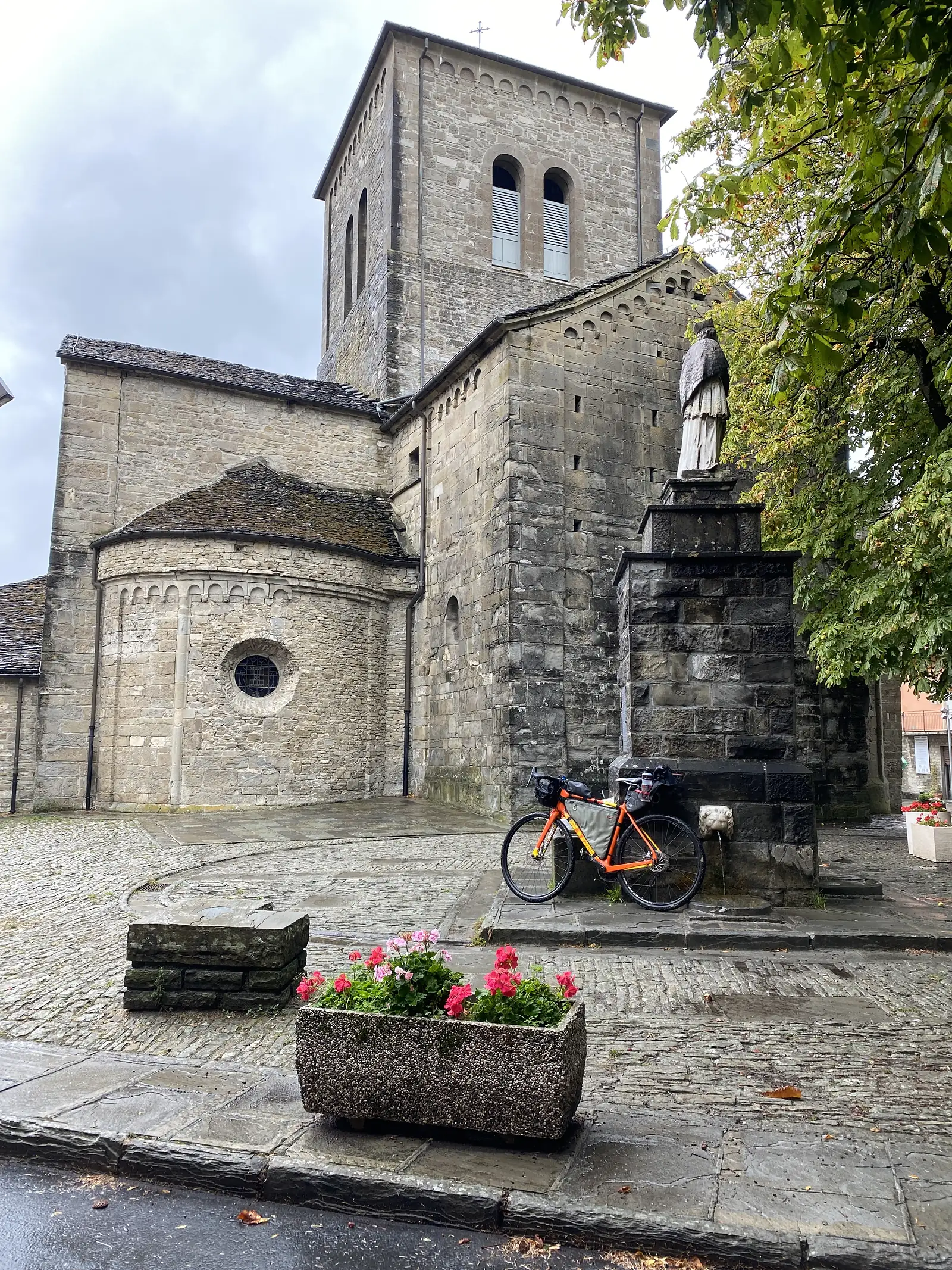 A loaded orange gravel bike leans against the stone base of a bell tower beside a wall fountain and pots of red geraniums in a village square.