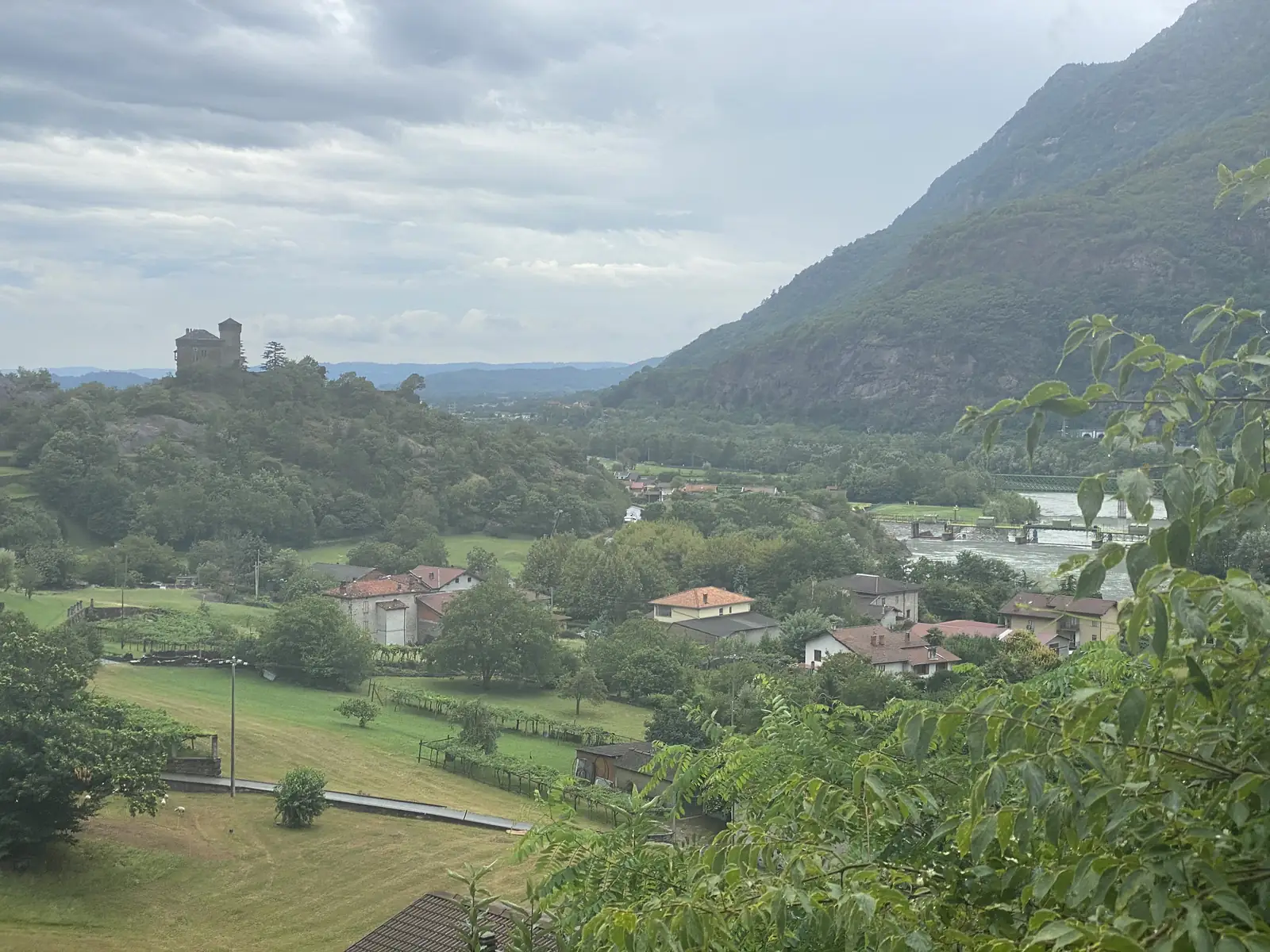 A crenellated stone castle crowns a wooded hilltop above a river valley, seen from a village garden with a small hilltop town visible in the distance.