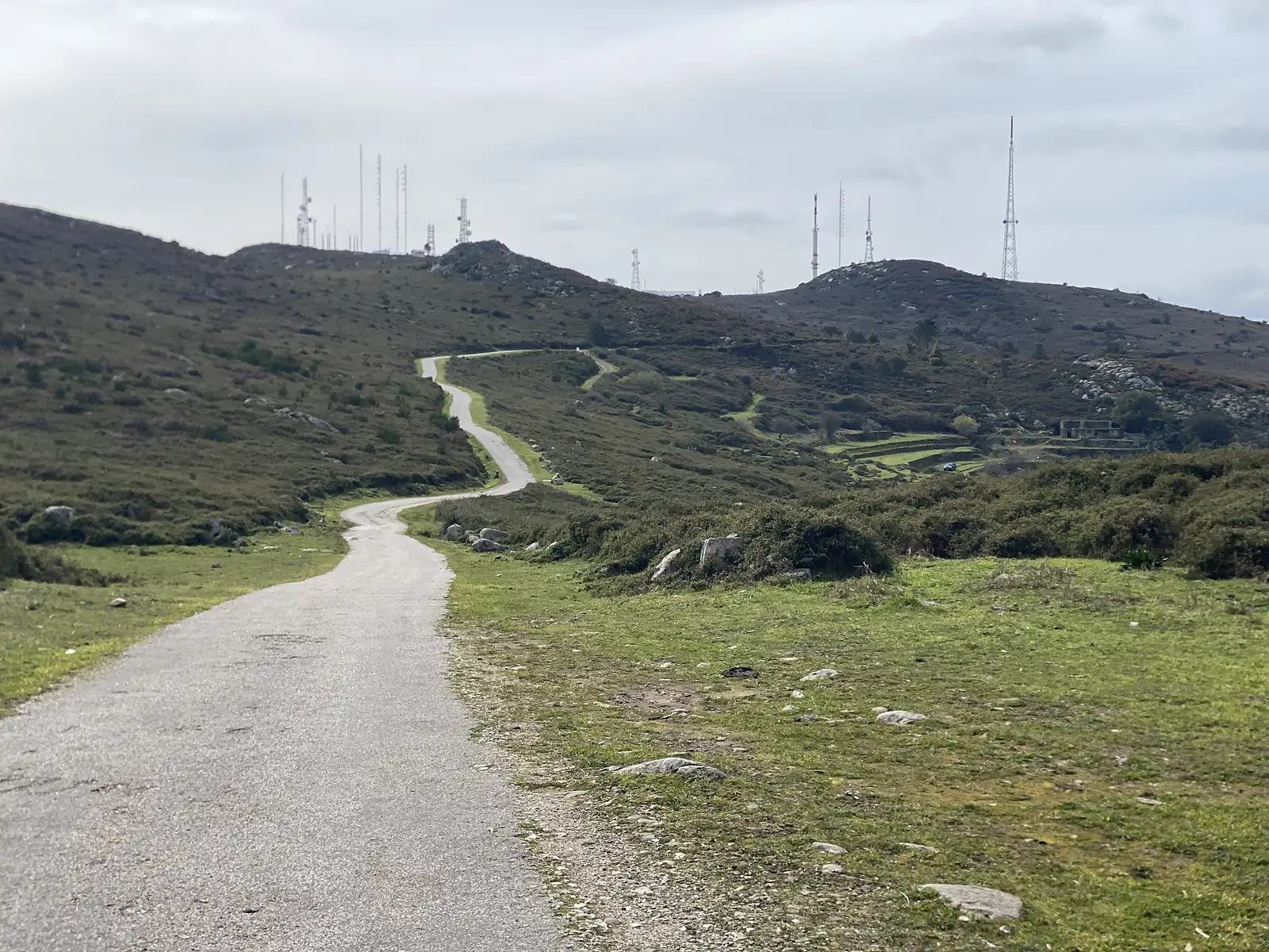 Narrow tarmac lane winding up over rough green moorland towards a summit bristling with telecommunications masts under a flat grey sky.