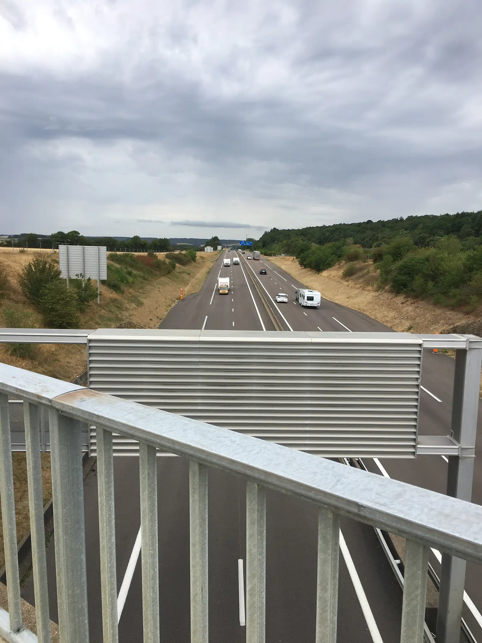 A view down from a motorway overpass onto a dual carriageway with a motorhome and cars passing below, wooded hills beyond.