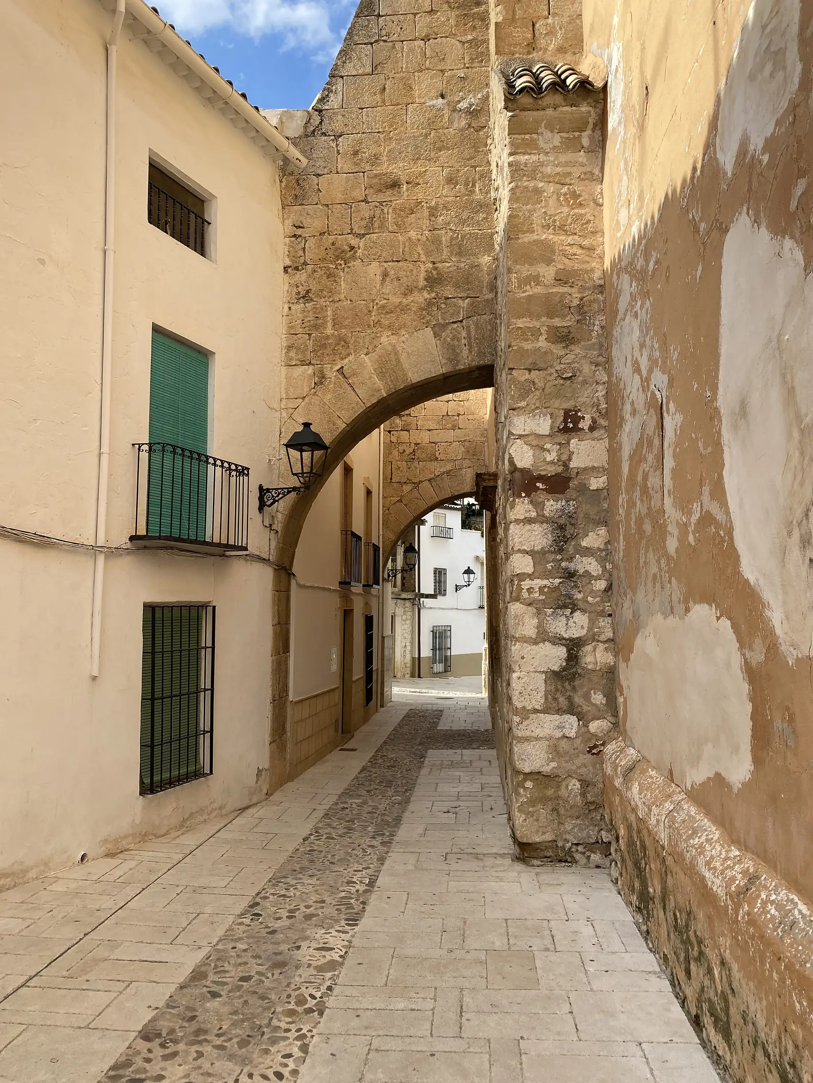 A paved lane passing beneath a massive stone arch buttress between an ochre wall and a house with a green-shuttered balcony.