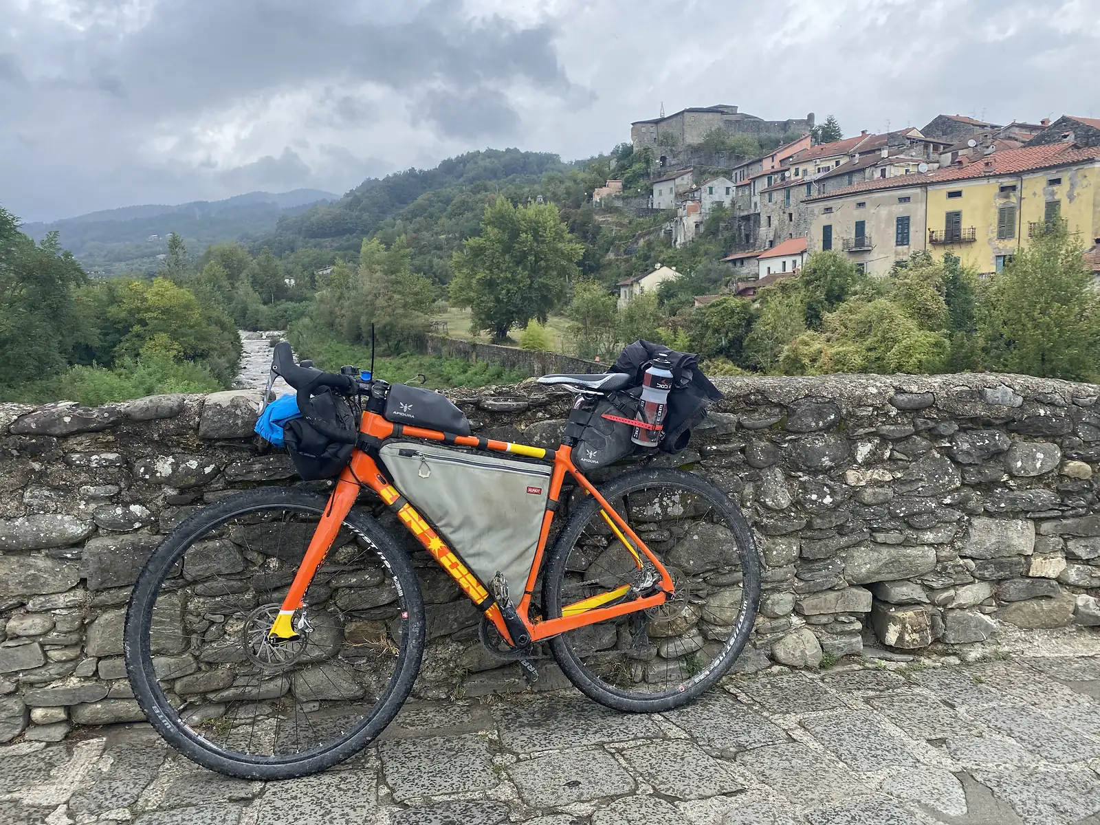 A loaded orange bikepacking bike leans on a stone parapet overlooking a river, with a hilltop town of stacked stone houses and a castle rising behind.