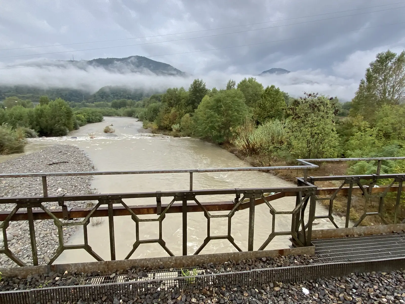 A riverside railway bridge with an ornate iron parapet overlooks a river running high and brown with floodwater, a gravel bar on one bank.