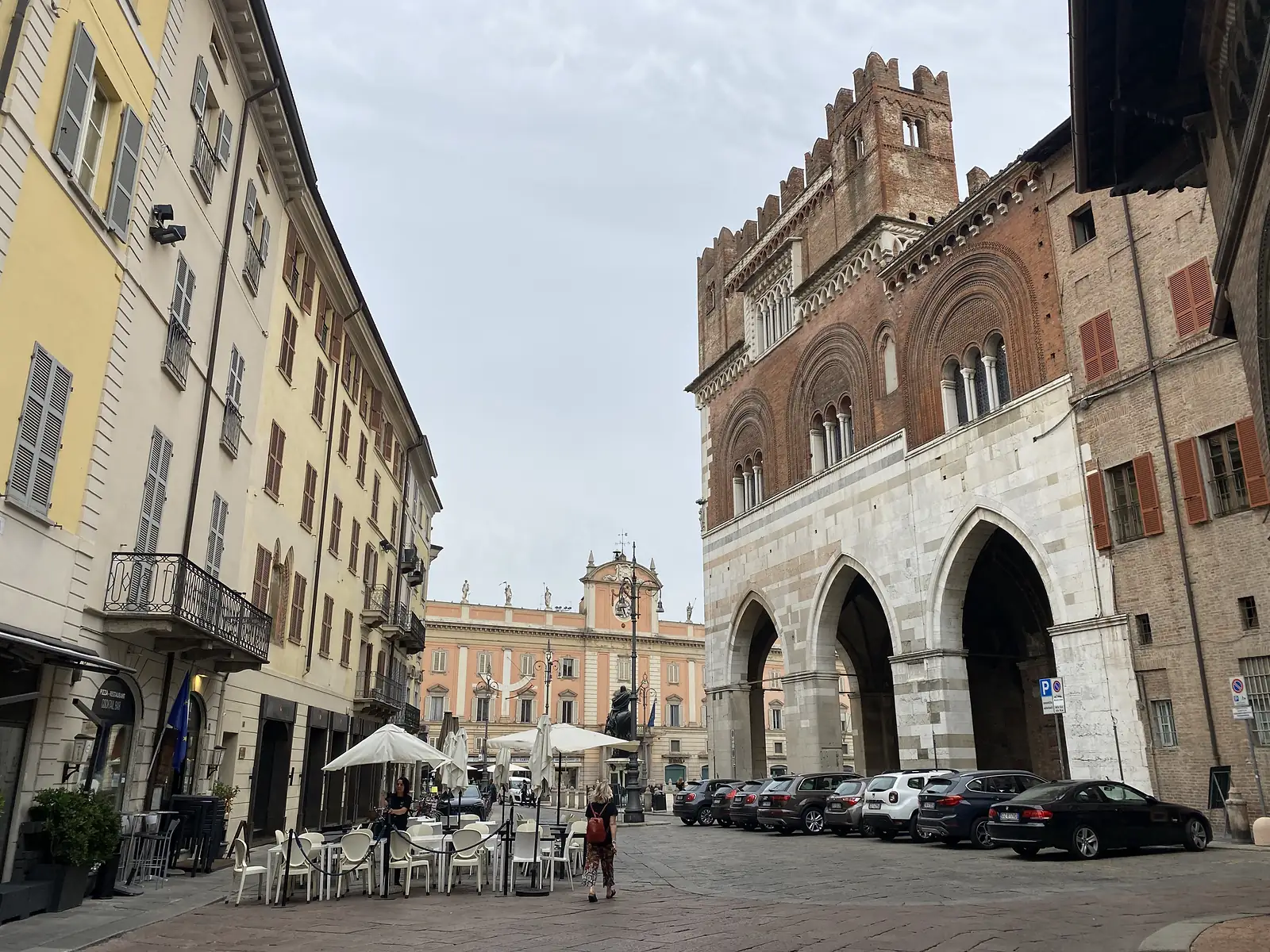 The fortified tower and Gothic arcade of the Palazzo Gotico rise above a Piacenza street, with cars parked along the piazza and a pale pink palazzo visible beyond.