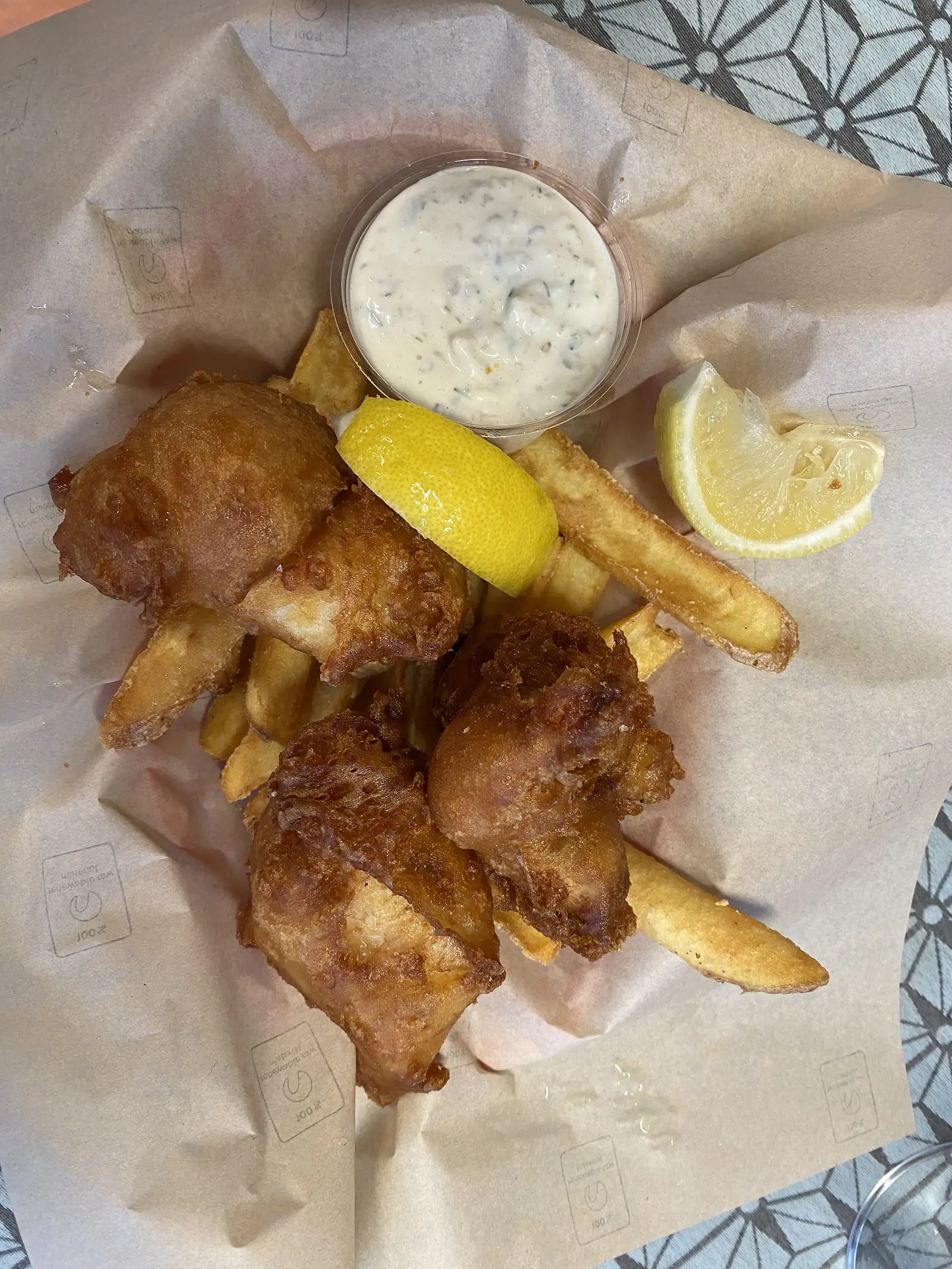 Battered fried fish and chips with a tub of tartare sauce and lemon wedges on greaseproof paper.
