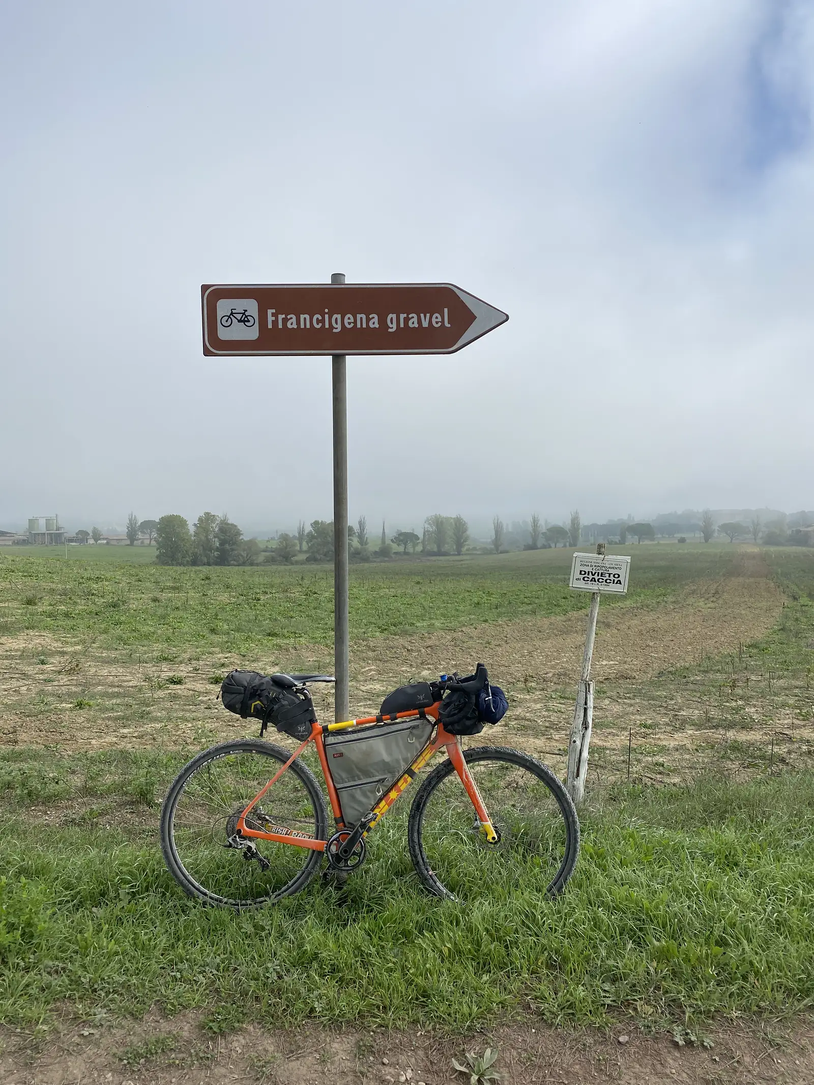 A loaded gravel bike propped beside a brown directional sign reading "Francigena gravel" with a cycling symbol, open farmland behind.