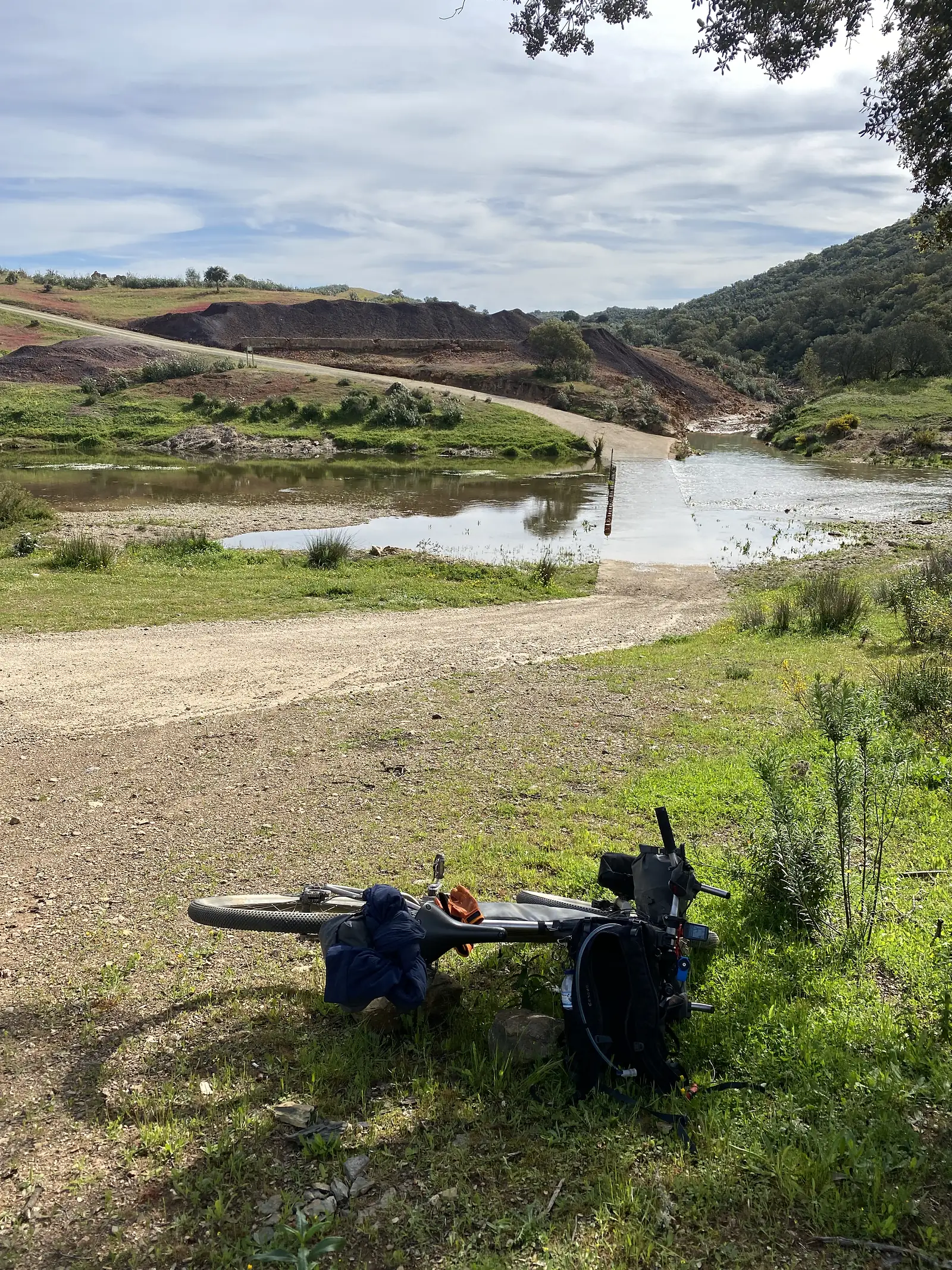 A loaded bikepacking bike laid on the grass beside a gravel track that drops to a concrete ford across a shallow brown river, with red-earth banks and green hills beyond.