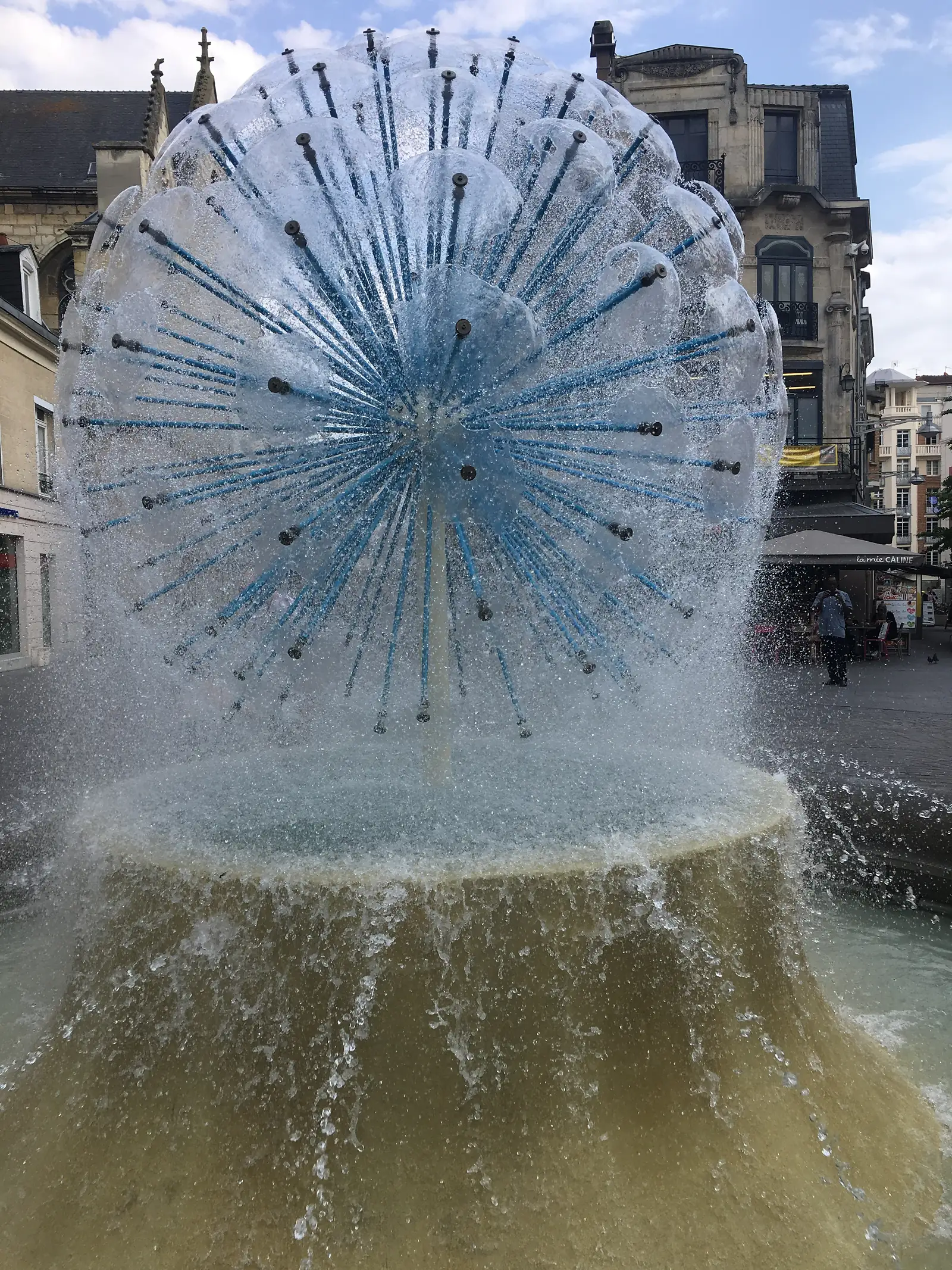 A large fountain sprays dozens of thin water jets in a fanned dome pattern in front of period townhouses on a French square.