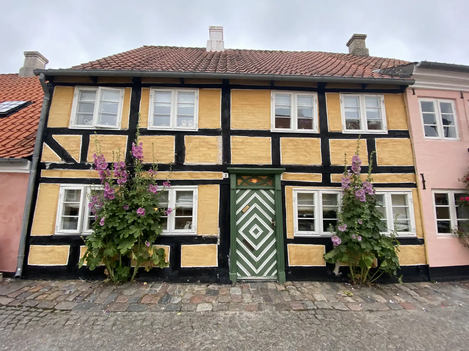 A yellow and black half-timbered house with a green-and-white chevron-painted door and hollyhocks in flower either side.
