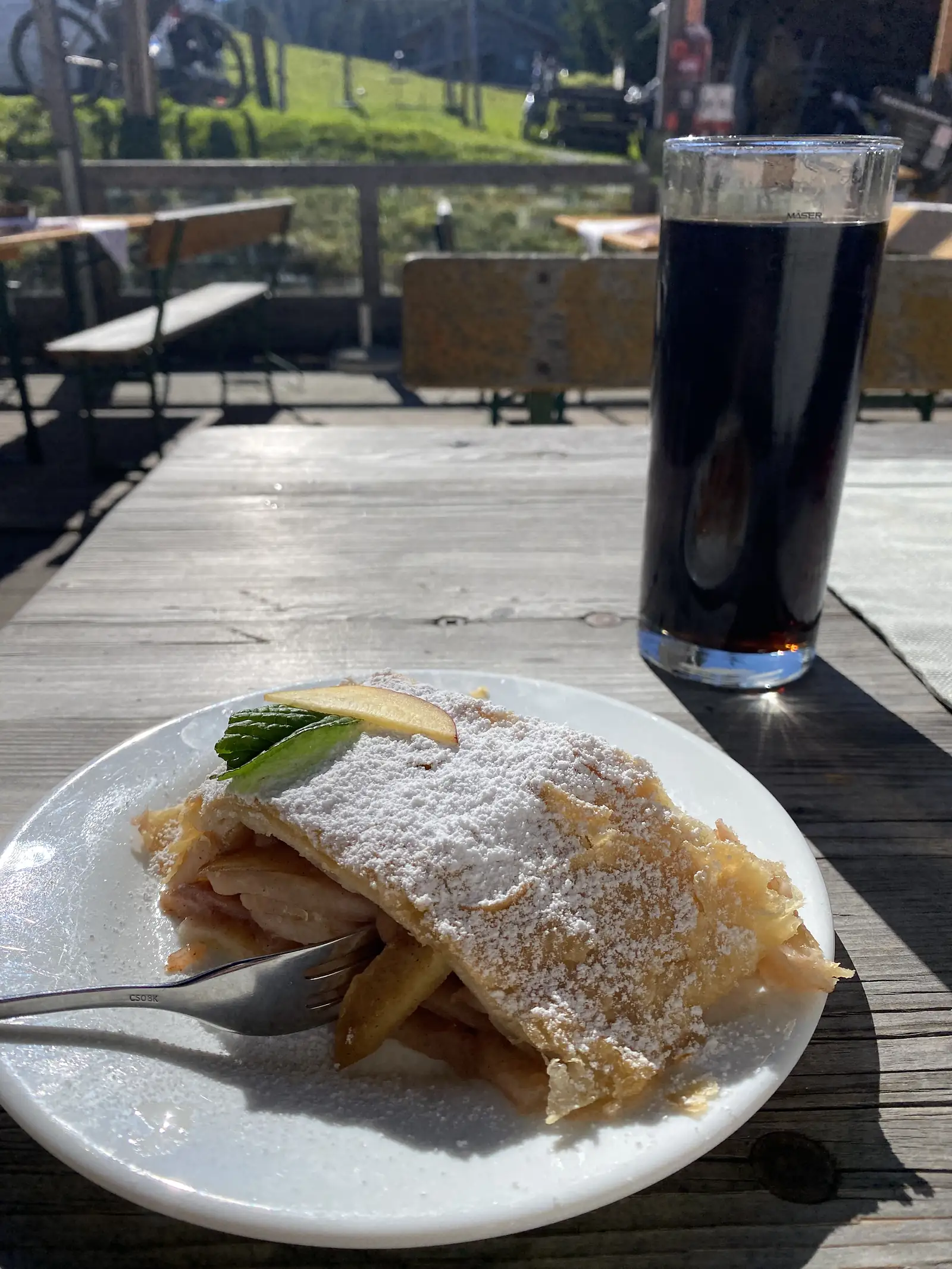 A slice of apple strudel dusted with icing sugar and a tall glass of dark beer on a sunlit wooden bench table, with bikes propped at the terrace rail behind.