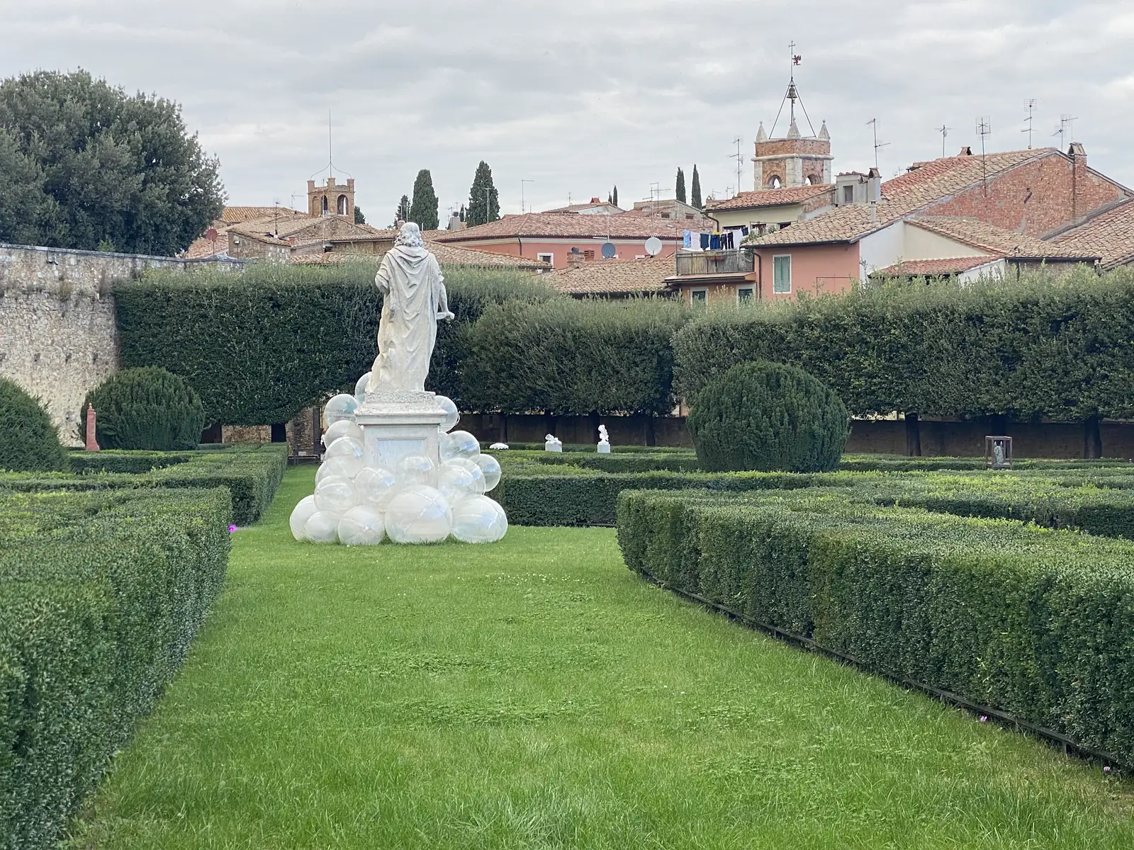 A classical robed statue on a plinth surrounded by large translucent glass spheres in a clipped box-hedge garden, with a hilltop town's rooftops and bell tower behind.