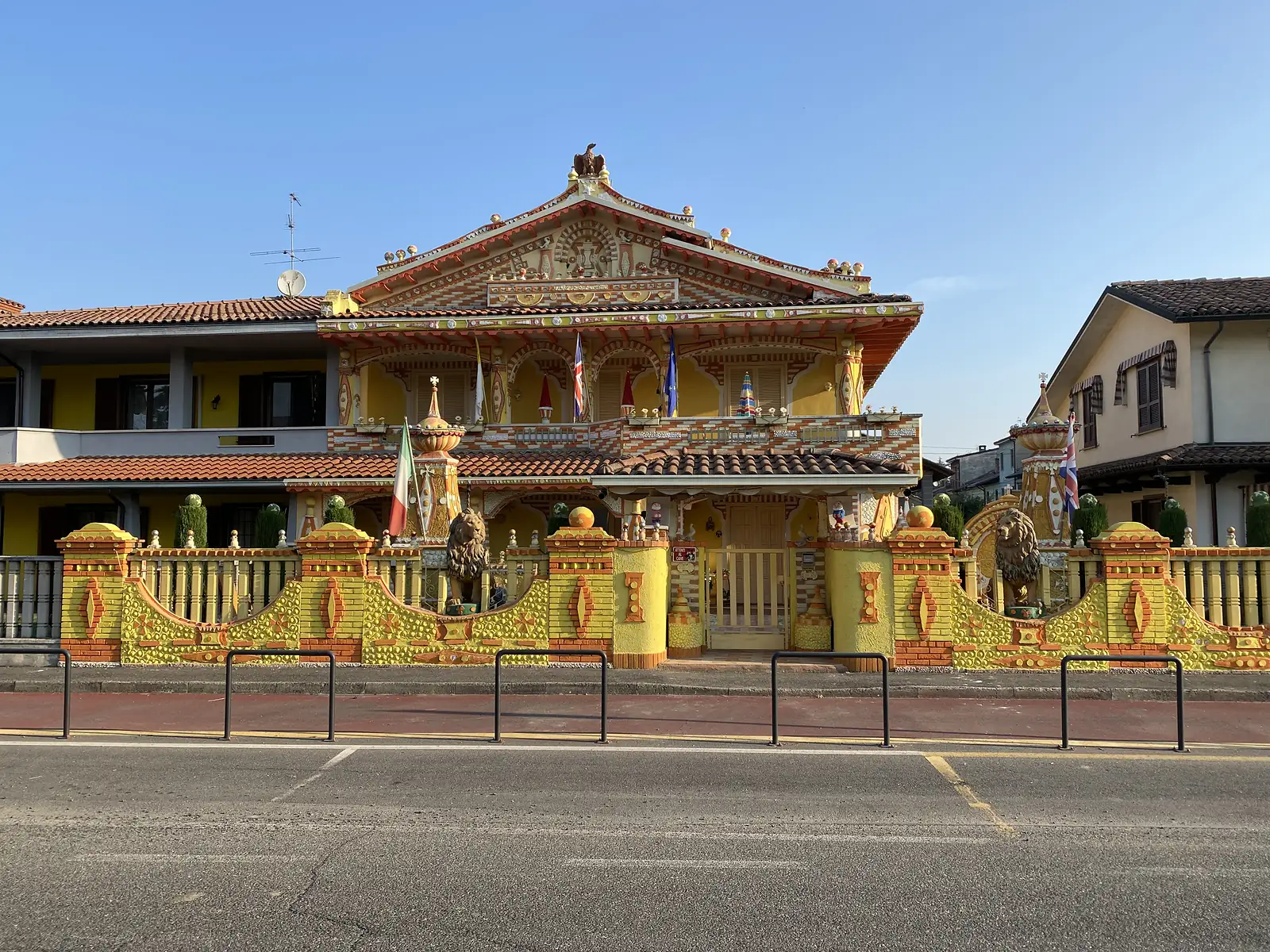 An elaborately decorated house painted yellow and white, with stone lion statues and an eagle finial atop its gateposts and flags flying from its balcony.