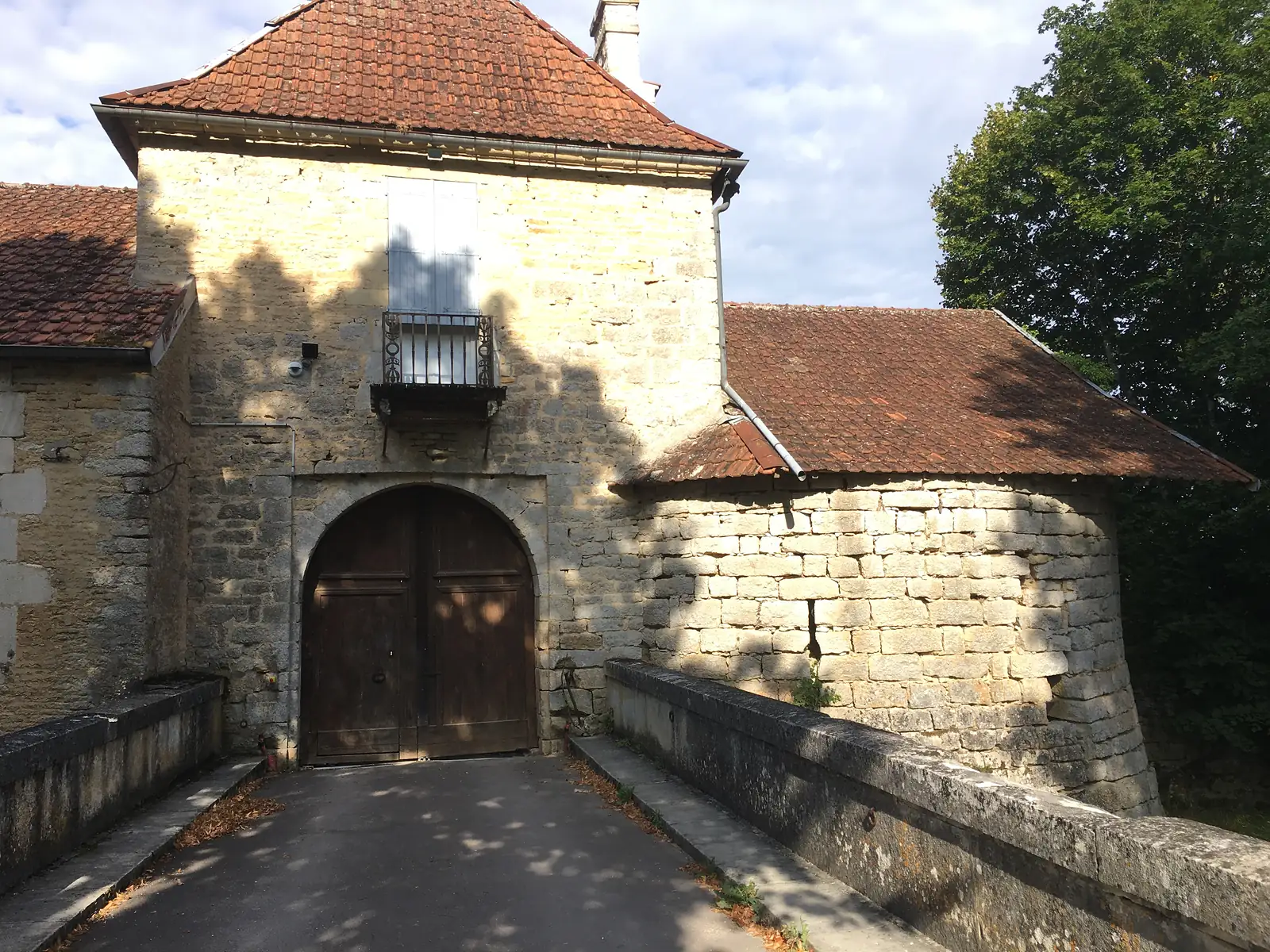 A stone gateway with an arched wooden door and shuttered window over a bridge, part of a fortified farmhouse or château entrance.