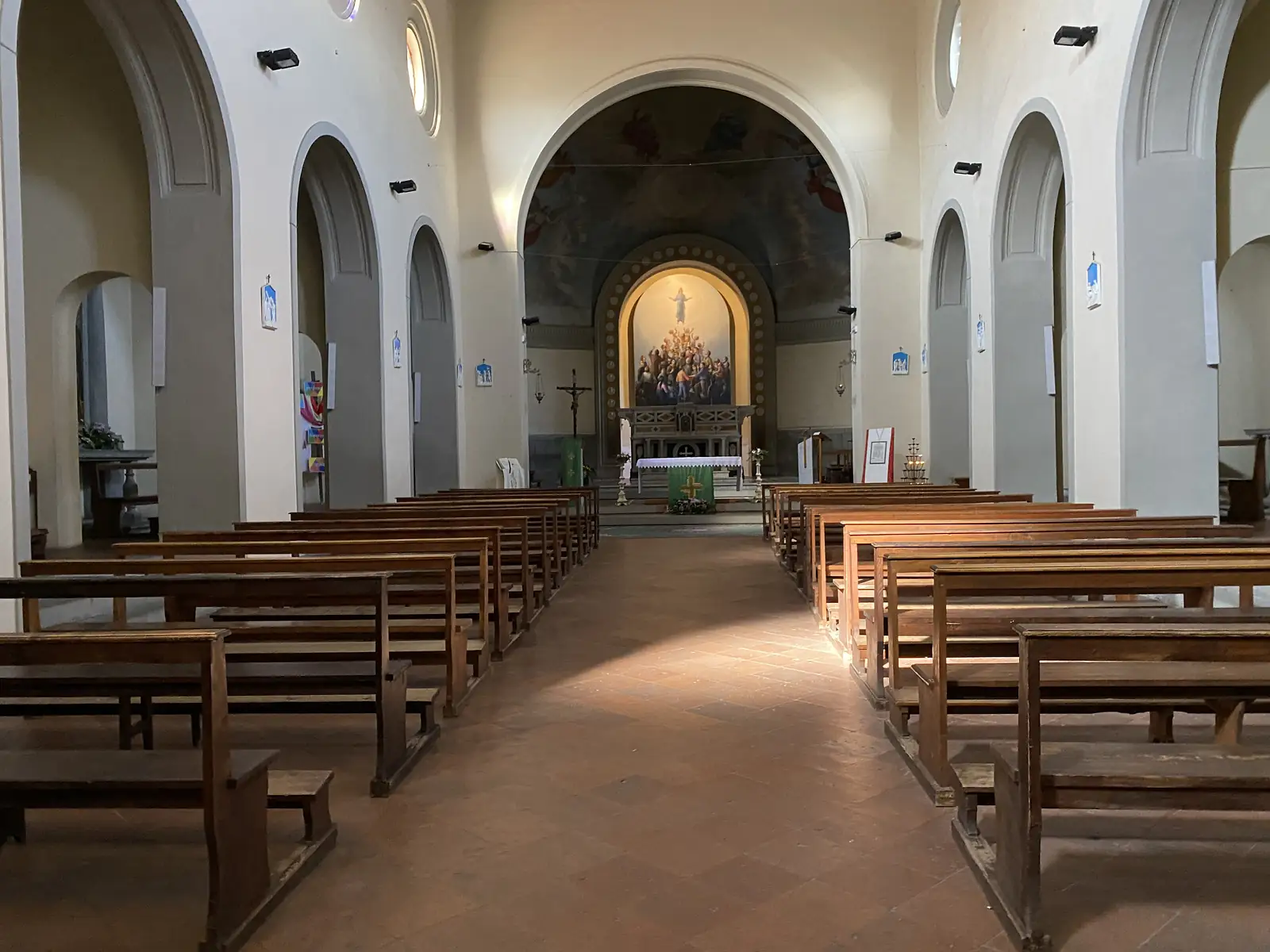 Empty wooden pews inside a stone church lead down the nave to an altar beneath a painted apse depicting a crowd gathered around a risen figure.