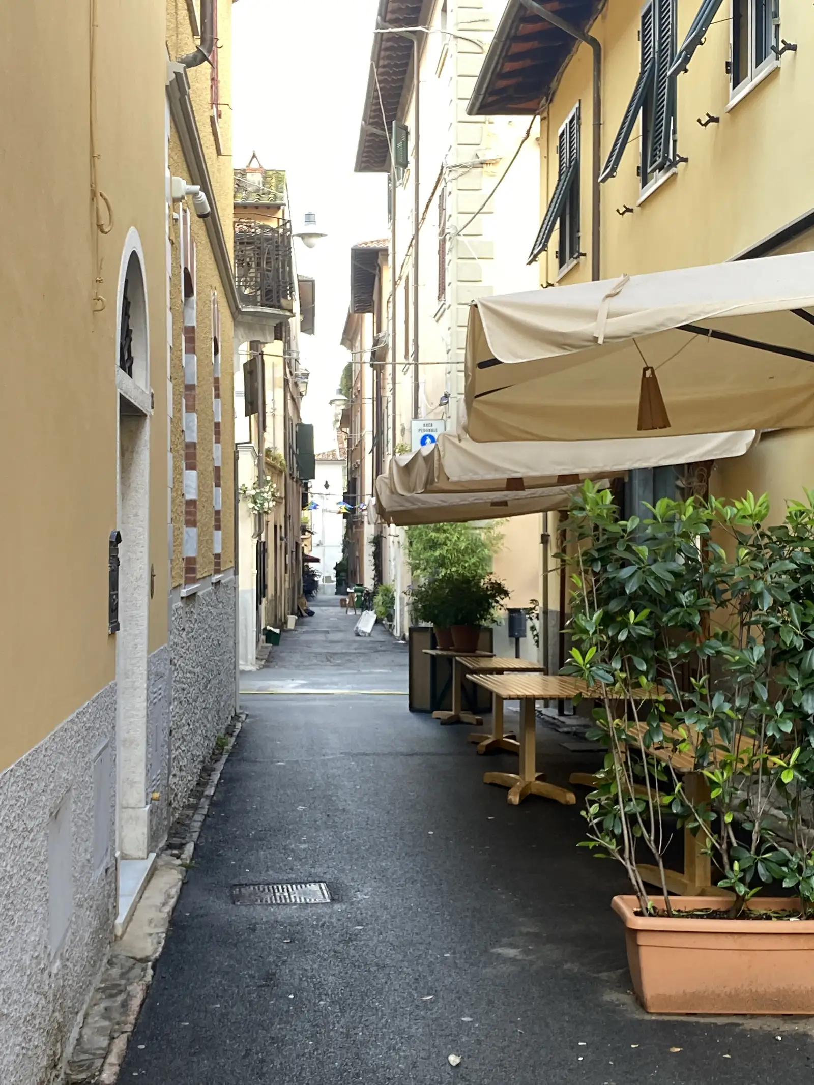 A shaded café terrace with folded parasols and potted shrubs sits beside a cobbled street marked with an 'Area Pedonale' pedestrian-zone sign.