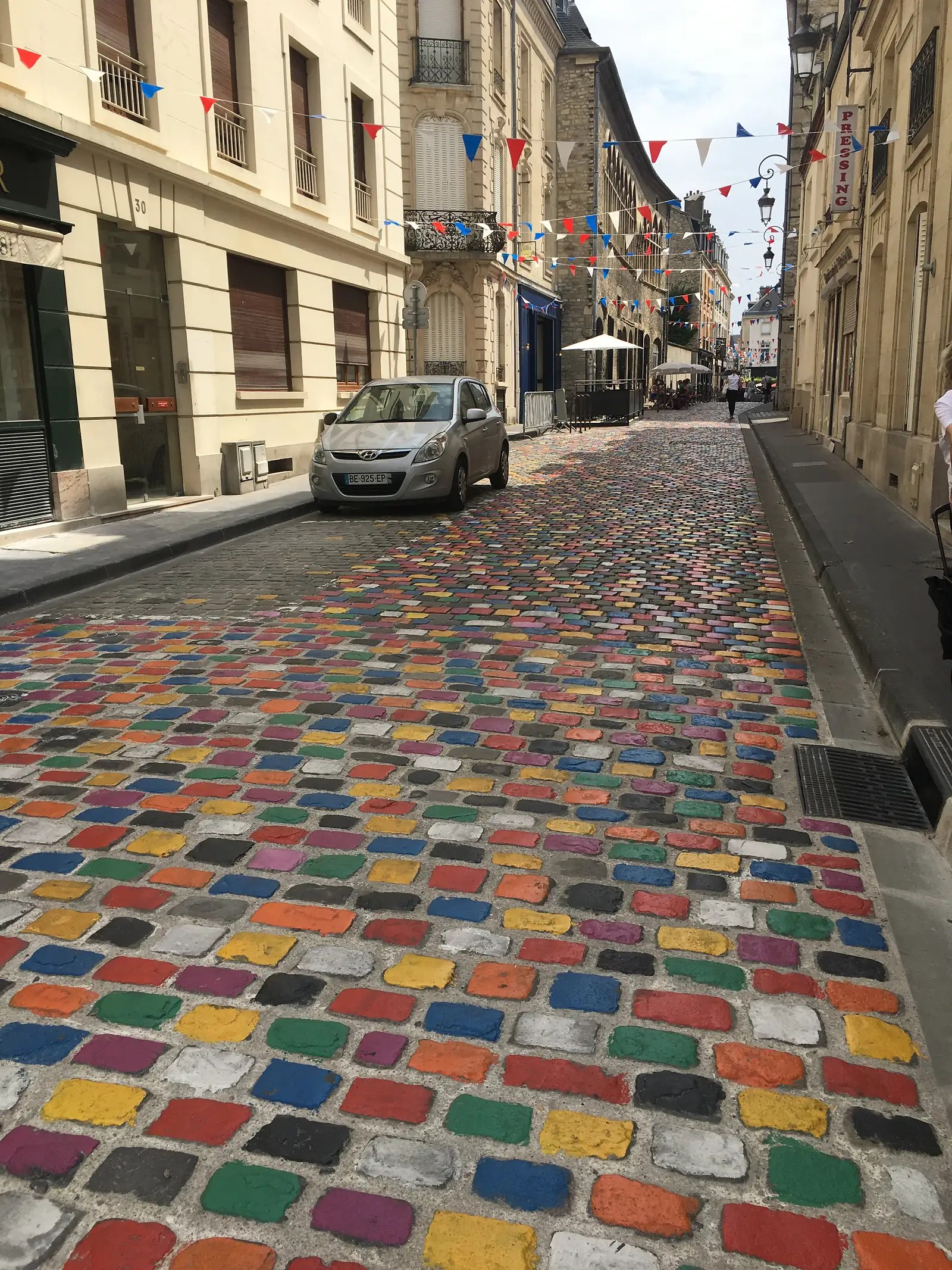 A cobbled street paved with stones hand-painted in a patchwork of bright colours leads towards a building strung with red, white and blue bunting.