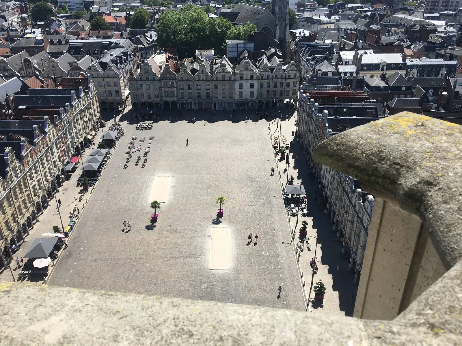 A large cobbled square lined on both sides by tall, gabled townhouses with ground-floor arcades, seen from high in a belfry with a weathered stone corner pinnacle in the foreground and a few people and potted palms scattered across the open flagstones below.