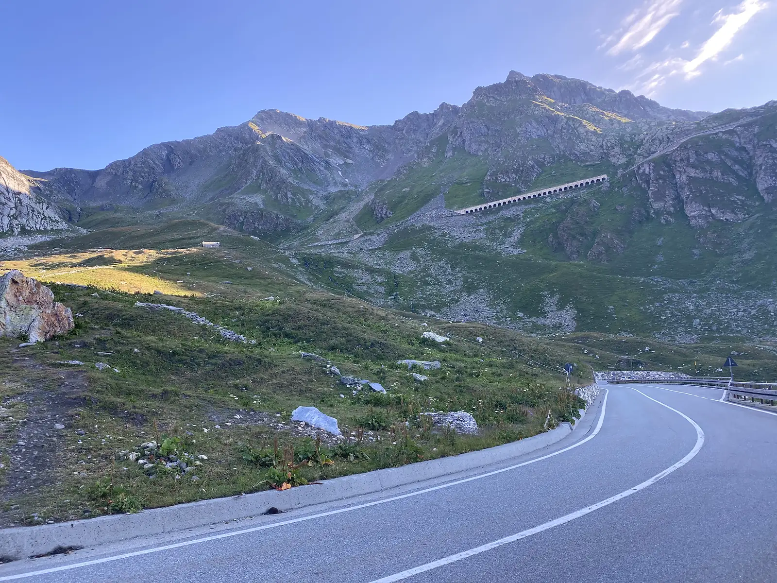 A tarmac road curves downhill below a rocky ridge near the Col du Grand-Saint-Bernard, with a long arched stone gallery built into the slope above and a small stone refuge in the valley beyond.
