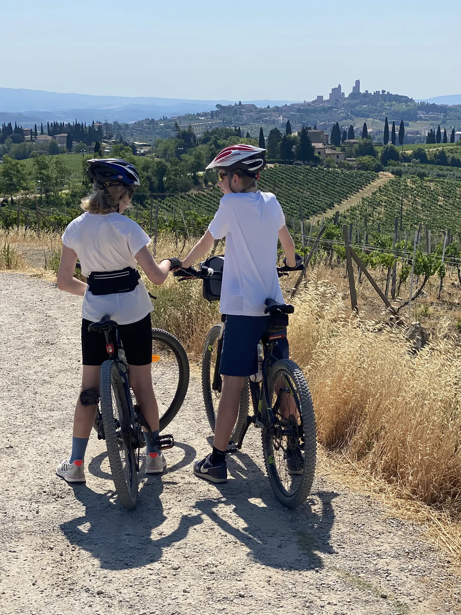 Two young cyclists with their bikes pause on a gravel track at golden hour, a towered hill town visible on the horizon behind them.