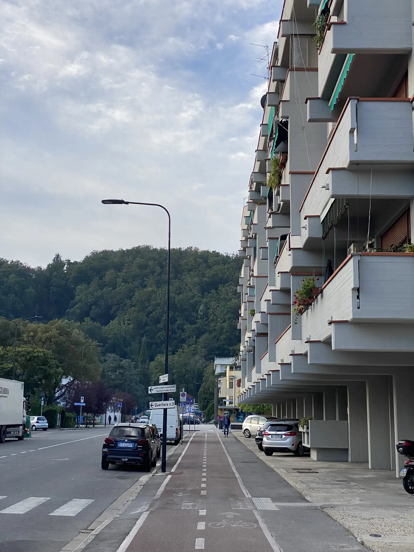 A suburban street lined with balconied apartment blocks and parked cars, with a signpost reading "Quartiere n.3" pointing along the road.