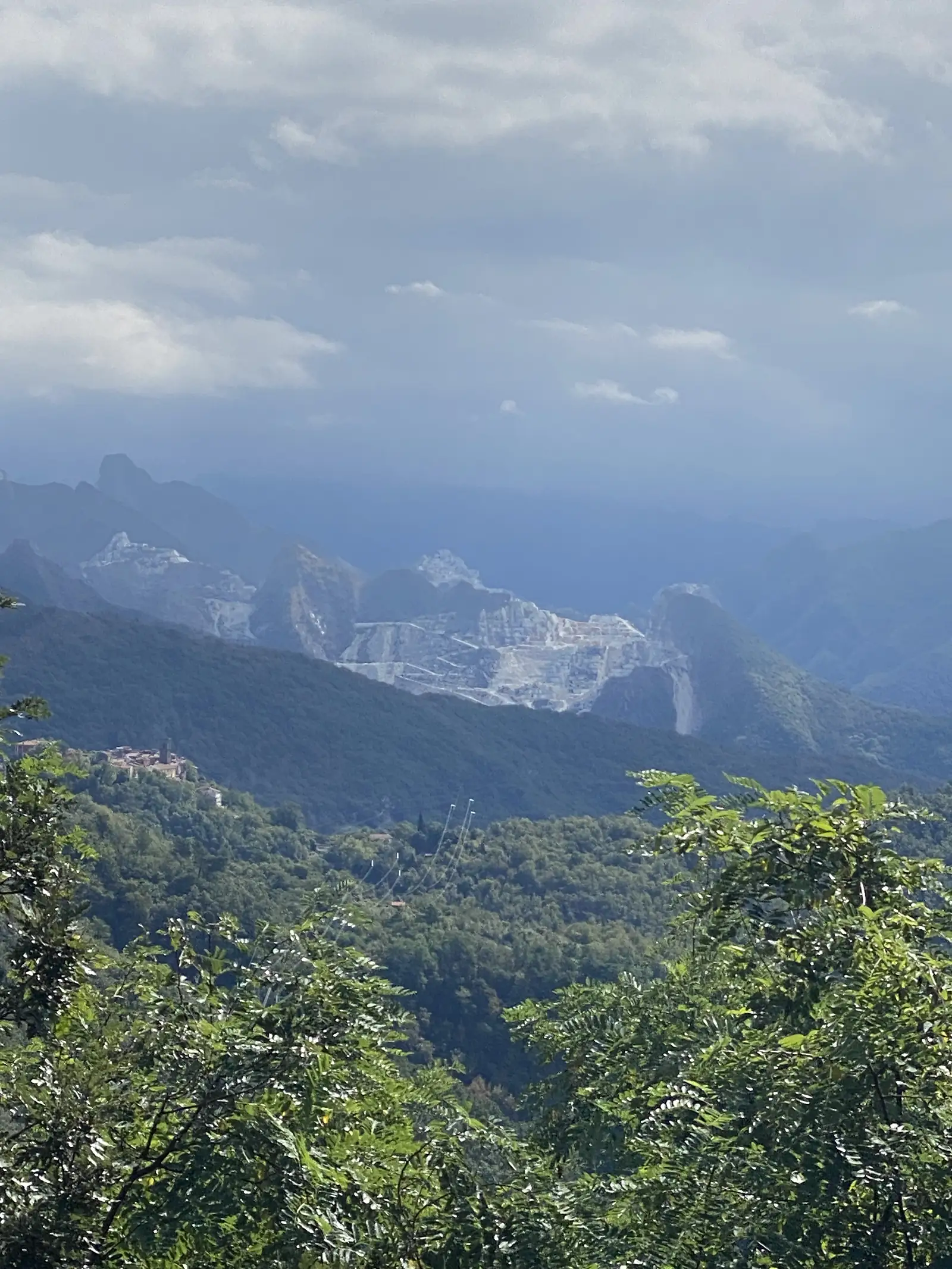 Pale marble quarry scars cutting into a green mountainside above a small village, seen across a wooded valley with pylon cables in the foreground.