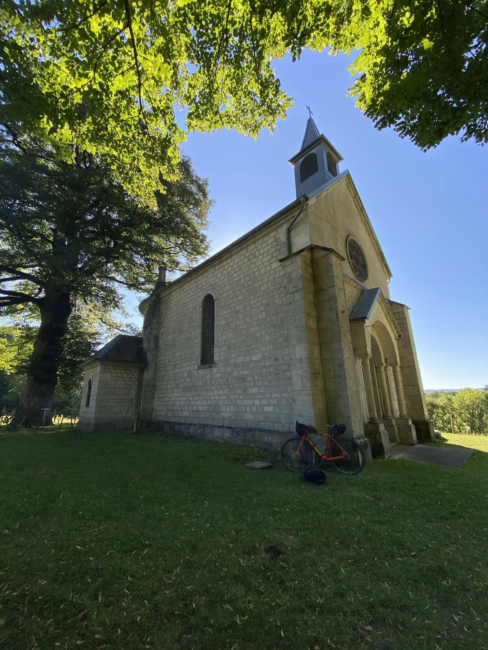 A loaded orange touring bike leans against the stone wall of a small chapel with a slate-roofed bell tower, its round window catching the sun.