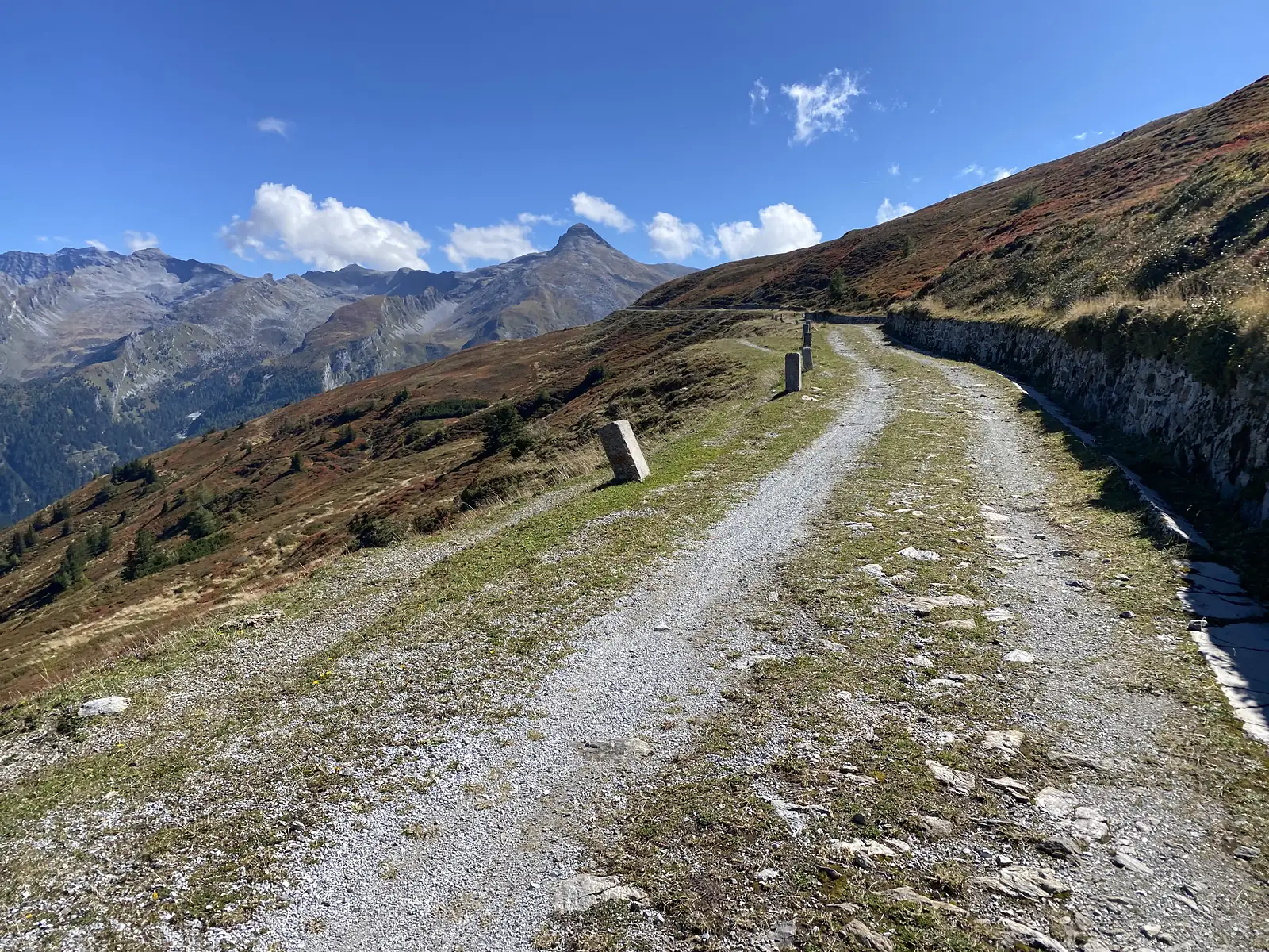 A rough double-track contours a steep russet hillside past stone marker posts, with a sharp pointed peak and further ranges ahead.