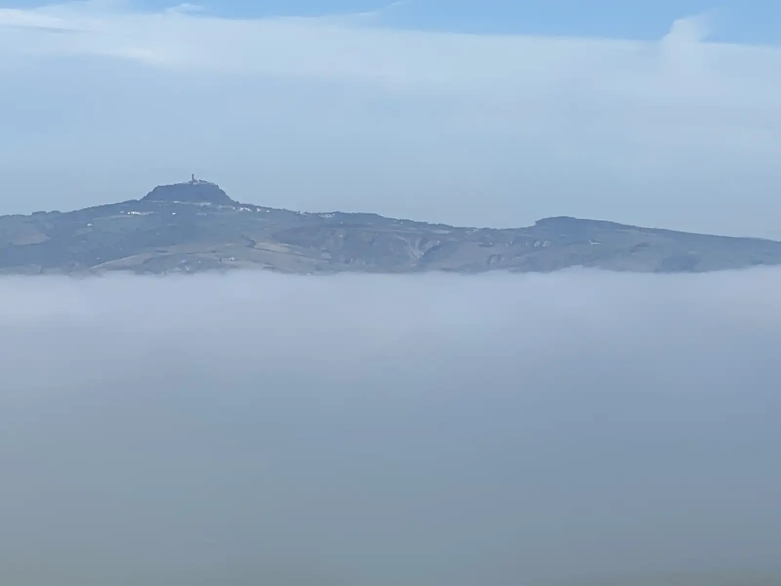 A hilltop village with a tower rises above a sea of low cloud filling the valley below.