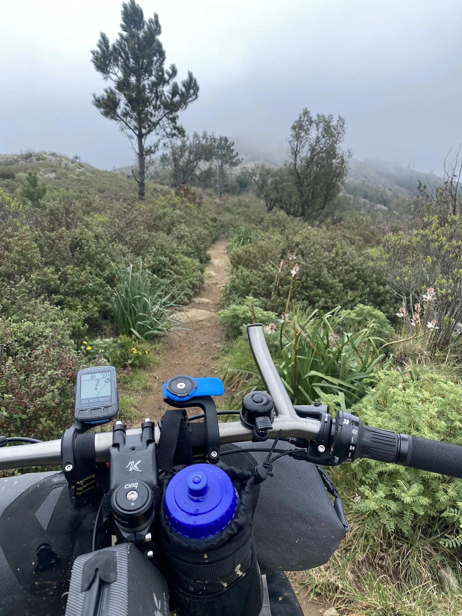 Rider's view over loaded handlebars, bottle and computer at the foot of a narrow singletrack climbing through heath towards a ridge lost in low cloud.