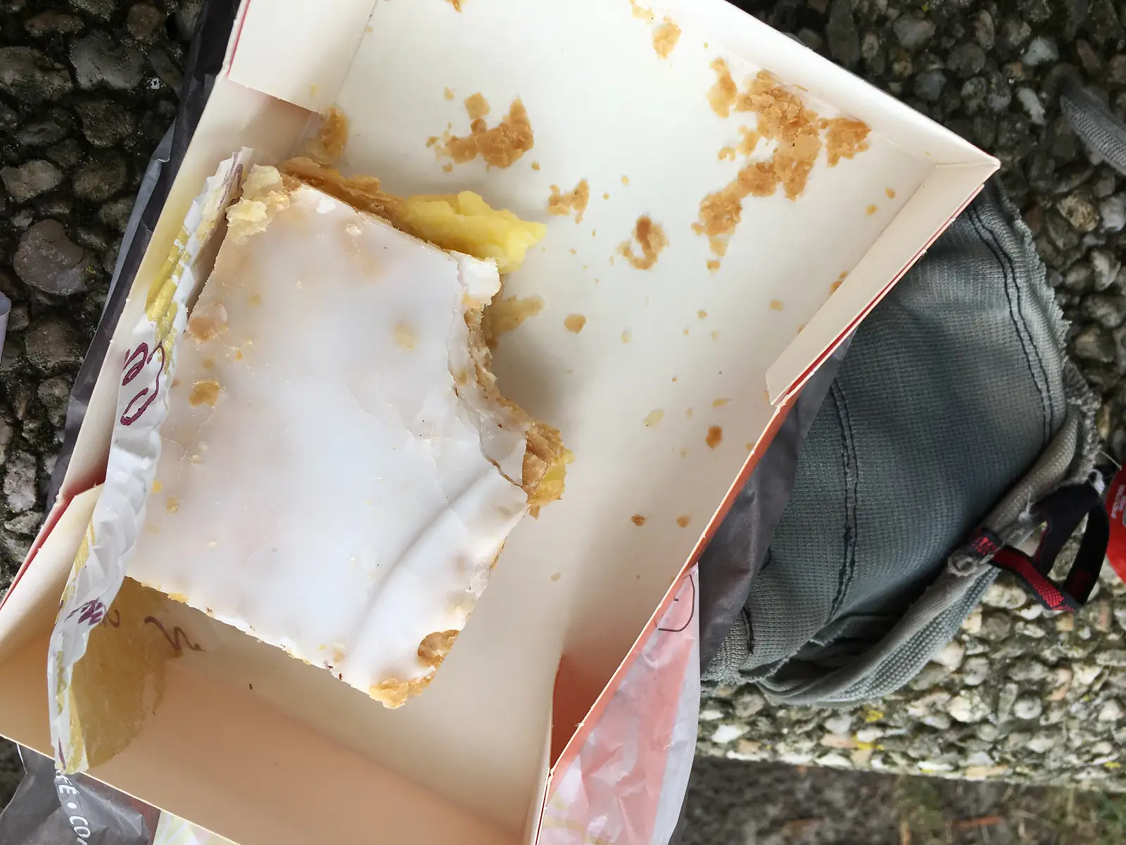 A half-eaten iced pastry, its yellow filling exposed, sits in an open takeaway box balanced on a rucksack.