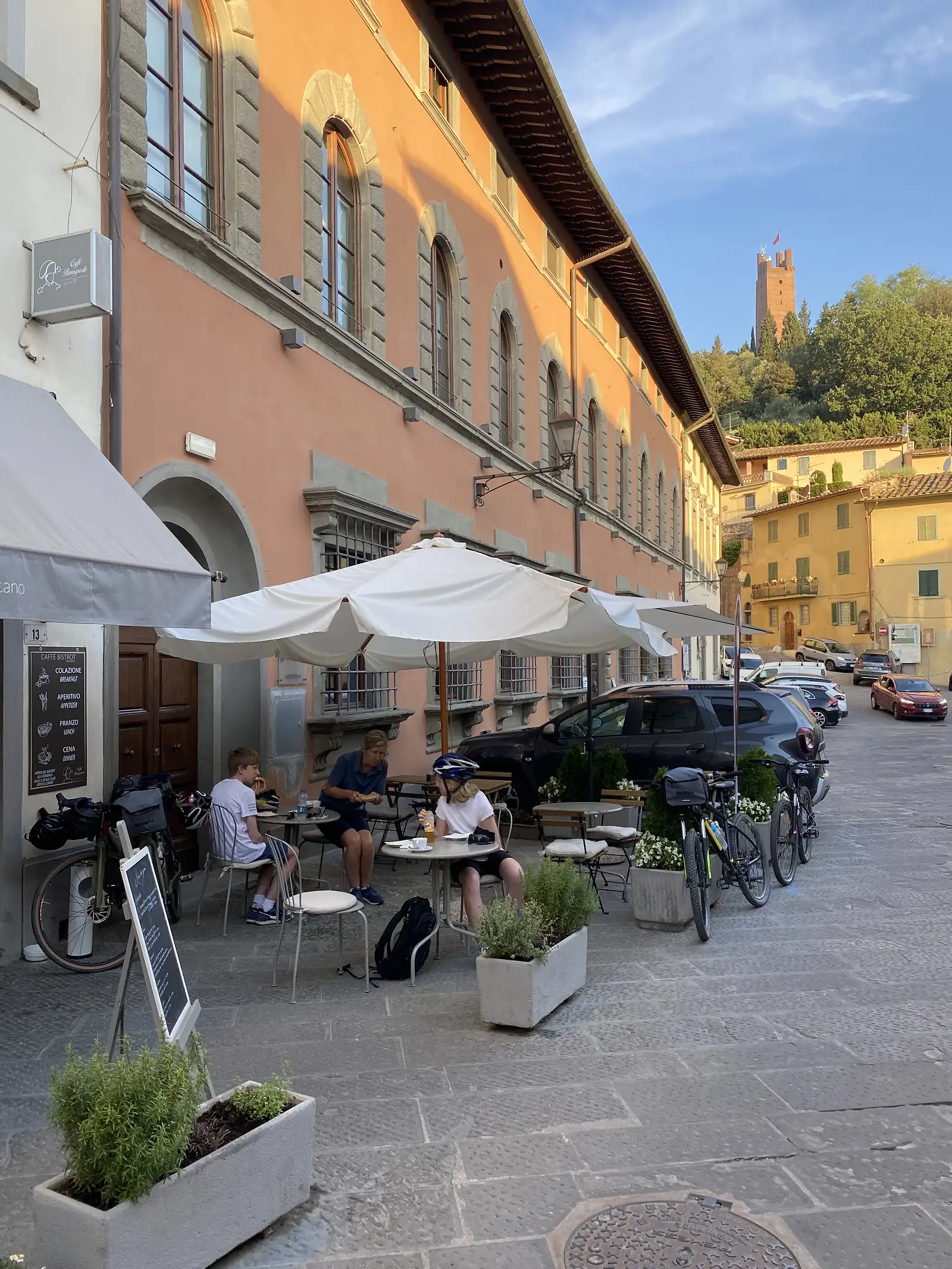 A family in cycling gear sits at an outdoor café table beneath a furled umbrella outside a "Caffè Bistrot" with a menu board, ochre-and-terracotta buildings lining the street and a distant tower visible under a blue sky.