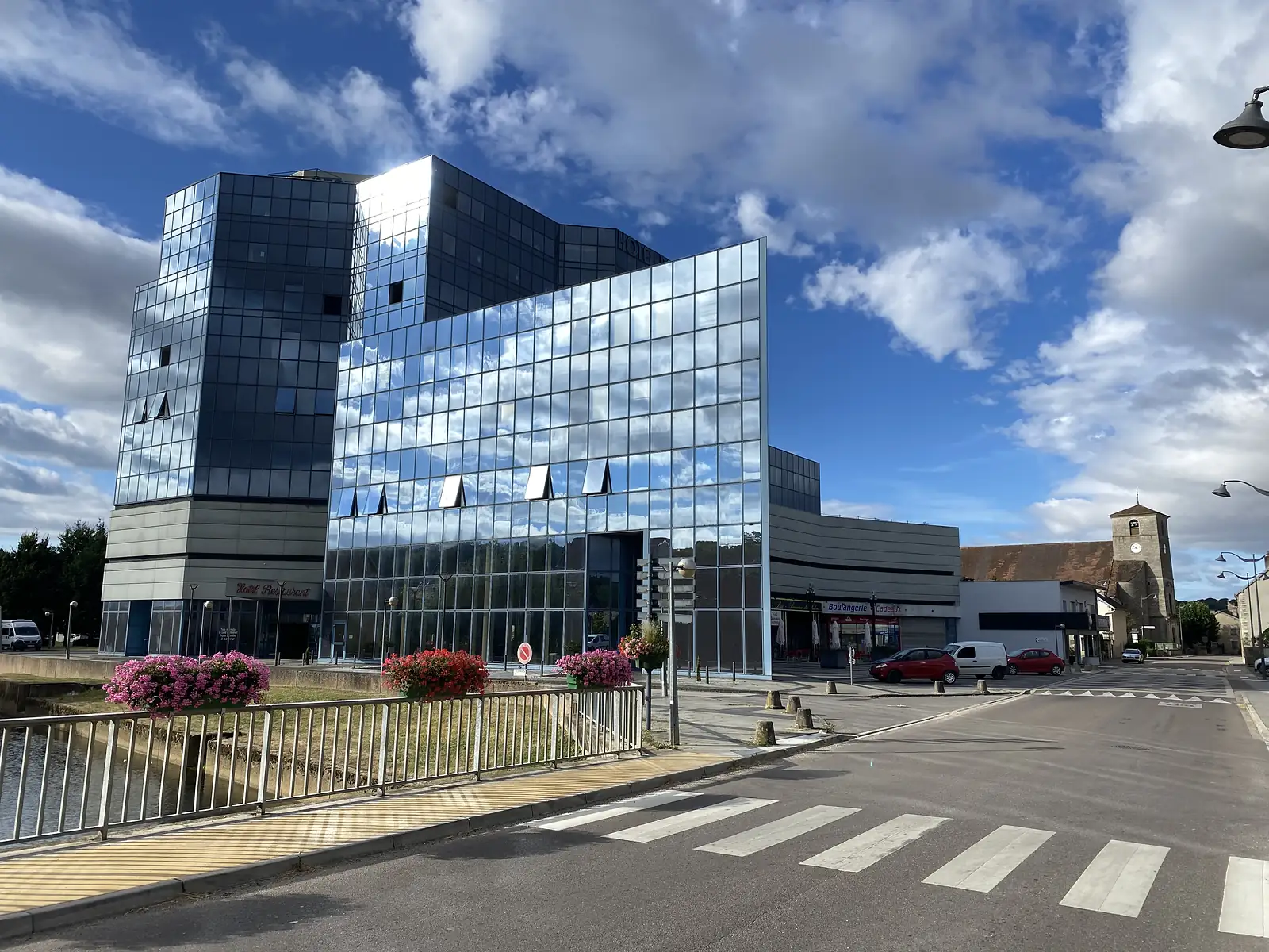 A mirrored-glass hotel building rises beside a canal, with a stone church tower and boulangerie visible further along the waterfront street.