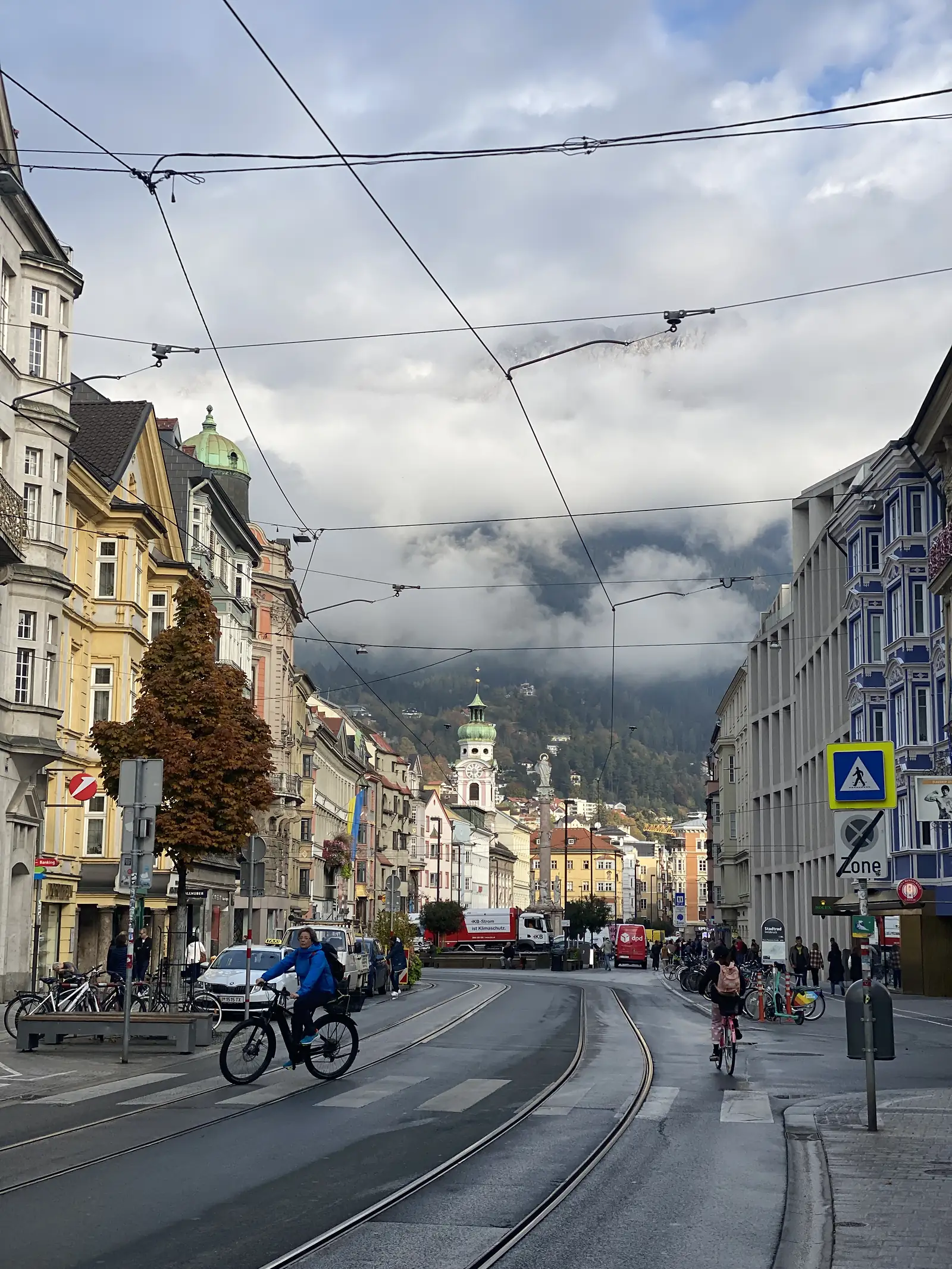 Pedestrians and cyclists pass along a street of colourful buildings in an alpine town, a 'Zone' pedestrian sign overhead and cloud-wreathed mountains behind.