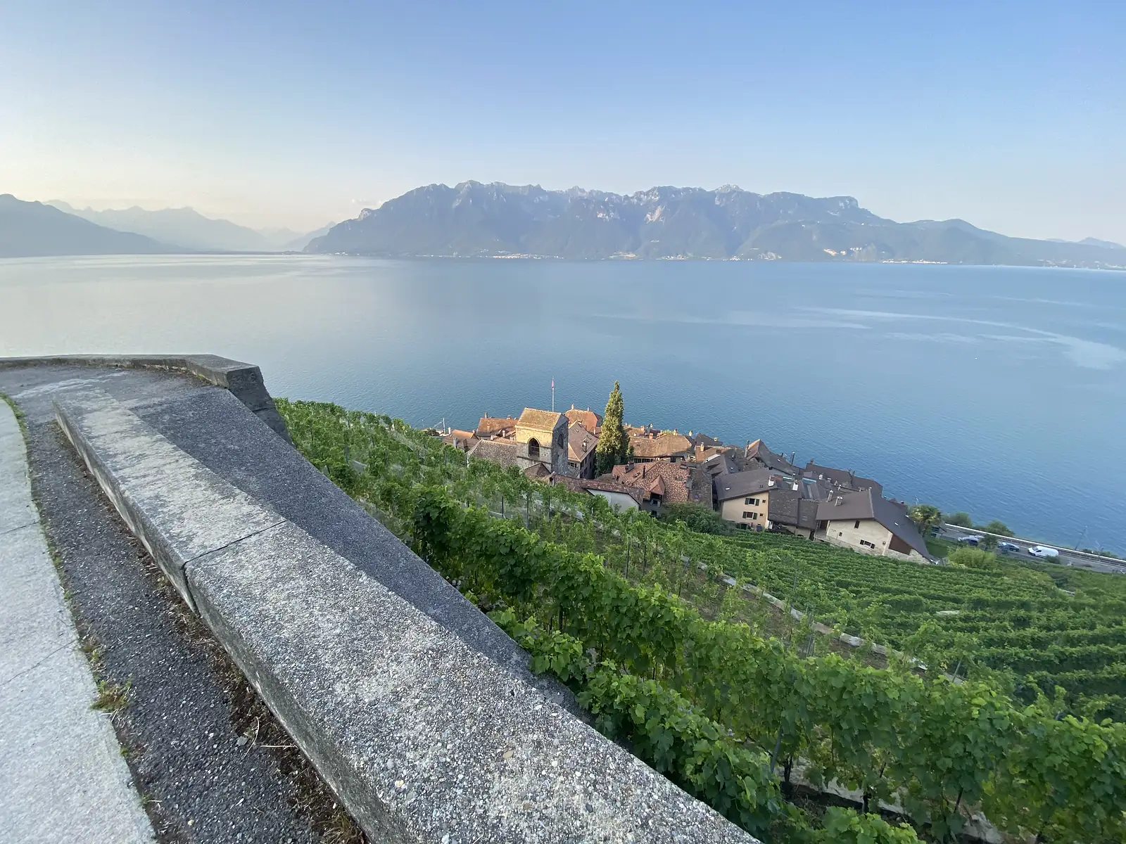 A low stone parapet above terraced lakeside vineyards leads the eye down to a cluster of red-roofed lakeshore buildings, with Lake Geneva and the far Alps beyond.