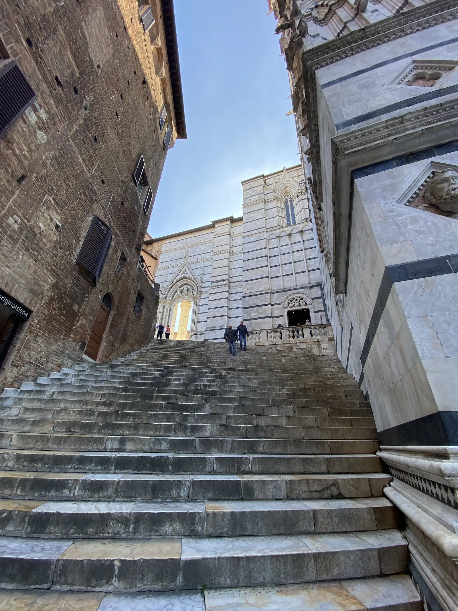 Looking up at the striped black-and-white marble facade and bell tower of a Gothic cathedral behind a brick building, with a shop sign reading 'originalsiena' confirming the location as Siena.