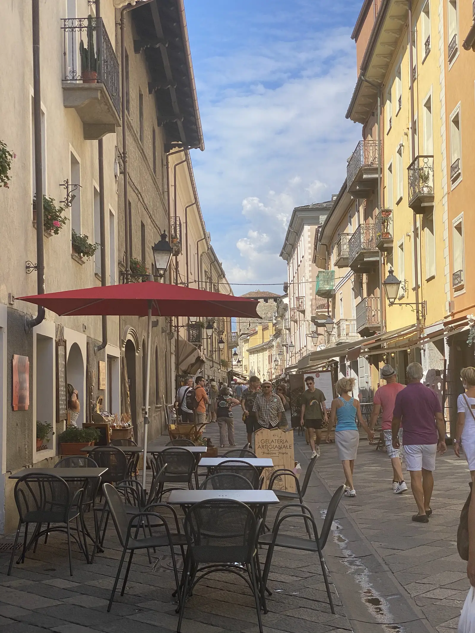 A café's red umbrella shades outdoor tables on a paved pedestrian street lined with pastel townhouses, shoppers and a wooden 'Gelateria Artigianale' sign visible further along.