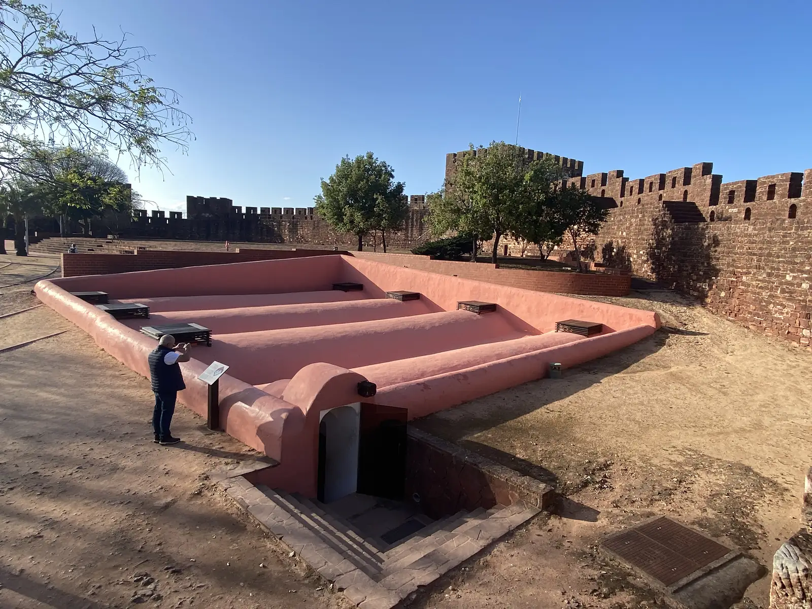 Restored terracotta-rendered cistern with a ridged, folded roof set below the crenellated red sandstone walls of a castle, a visitor in a sun hat reading the information board beside it.