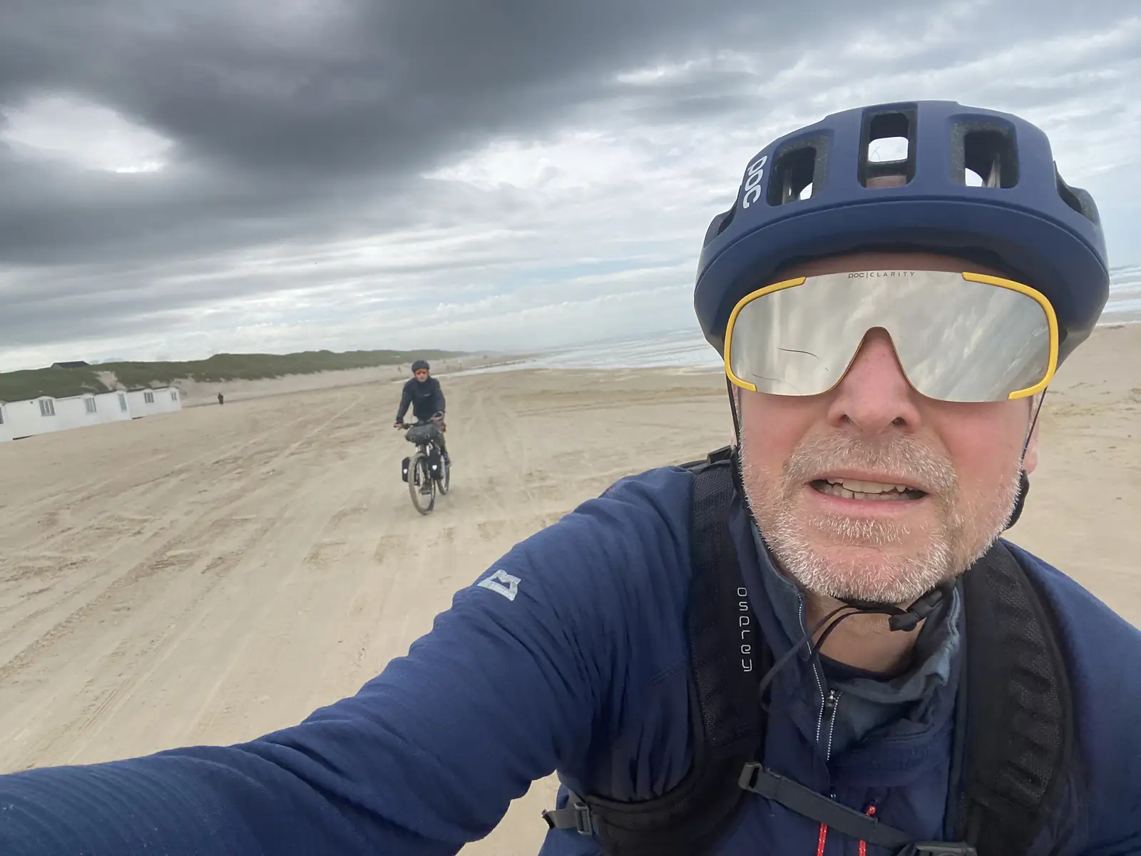 A close-up selfie of a cyclist in a blue helmet and mirrored sunglasses riding along a wide sandy beach, with a second rider following and beach huts on the dunes.