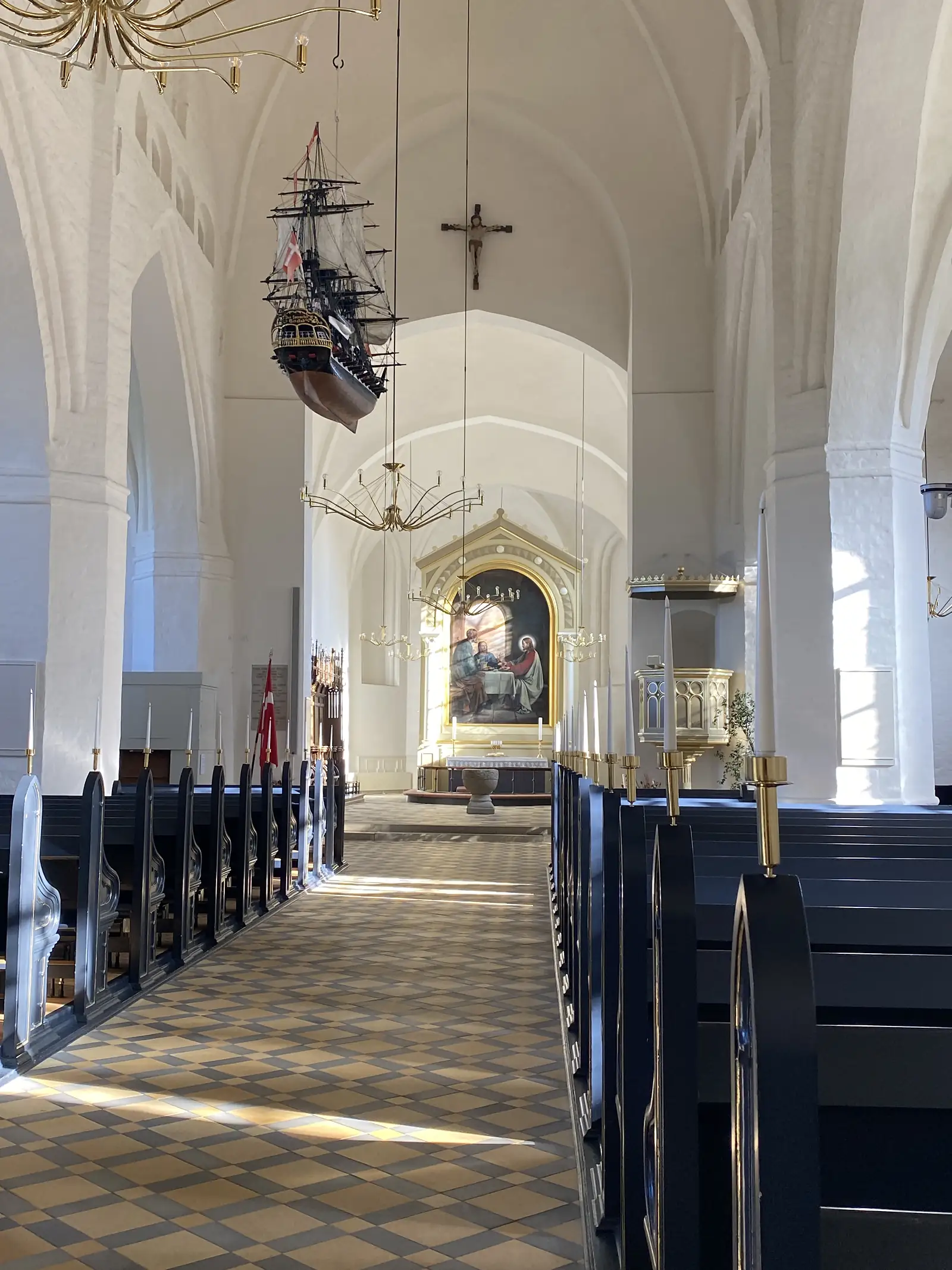 The whitewashed vaulted interior of a church with a model sailing ship hung from the ceiling above dark pews and brass chandeliers.