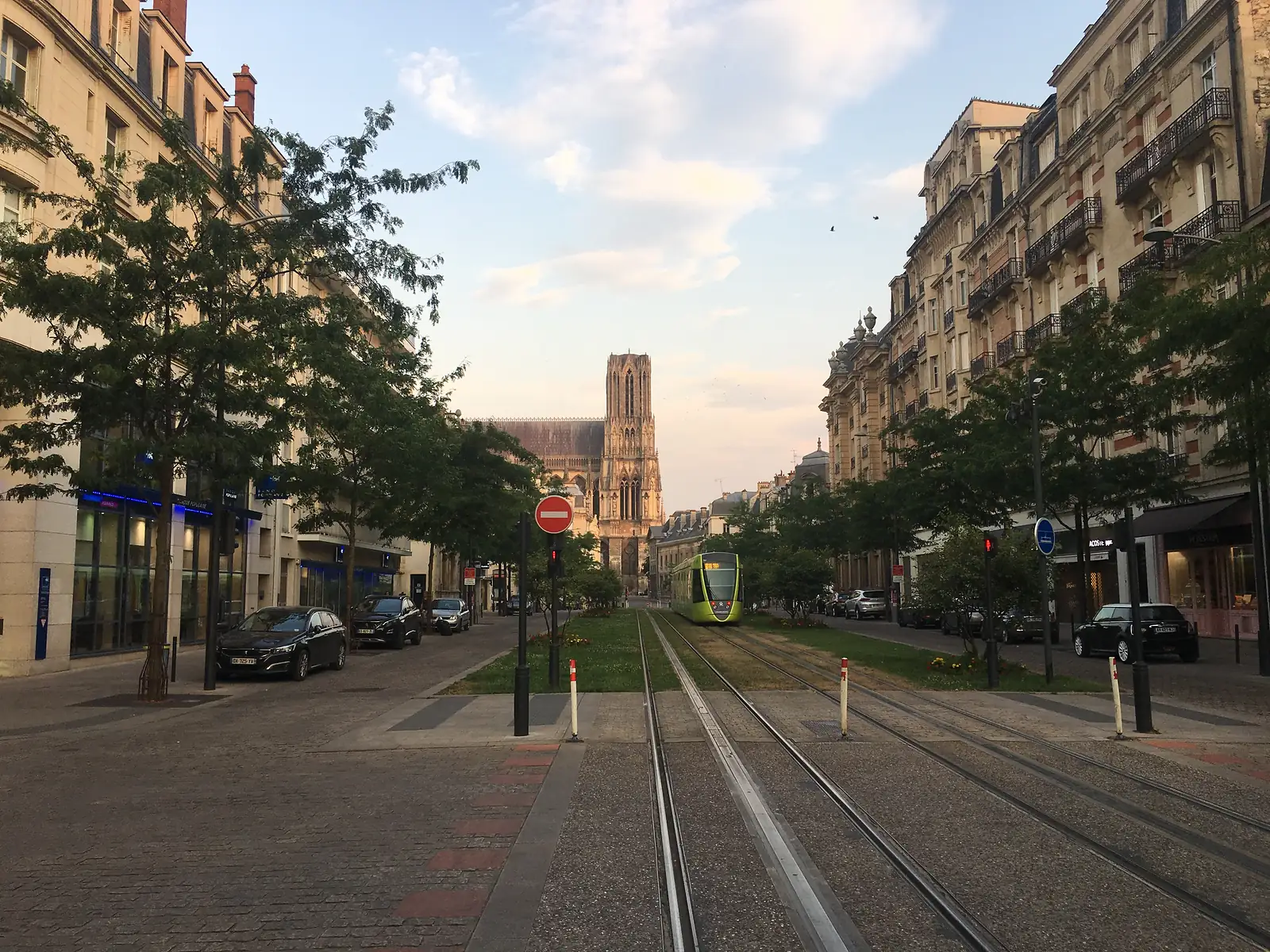 A green tram runs along grassy tracks down a tree-lined boulevard towards the twin towers of Reims cathedral glowing in evening light.
