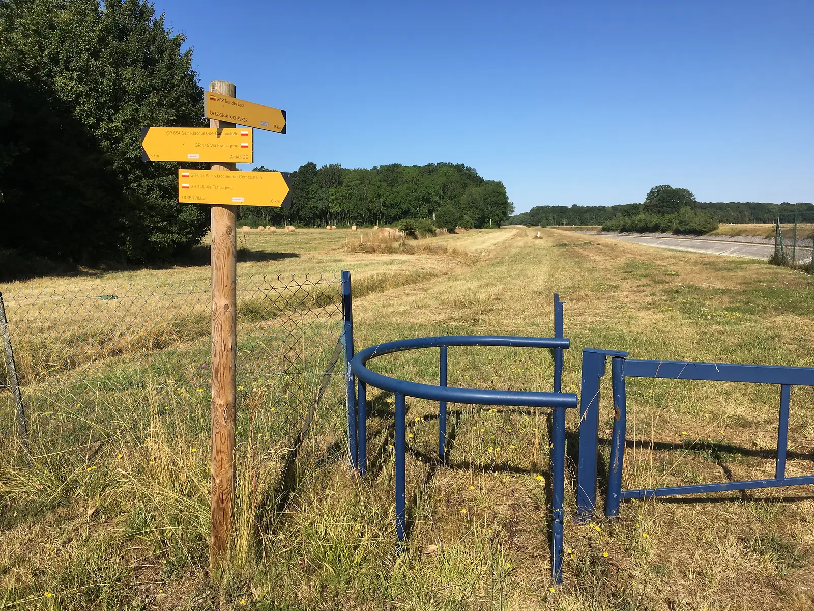 A cluster of yellow waymark signs for the GR654 Saint-Jacques-de-Compostelle and GR145 Via Francigena routes on a wooden post beside a field of hay bales, with a broken blue metal gate nearby.