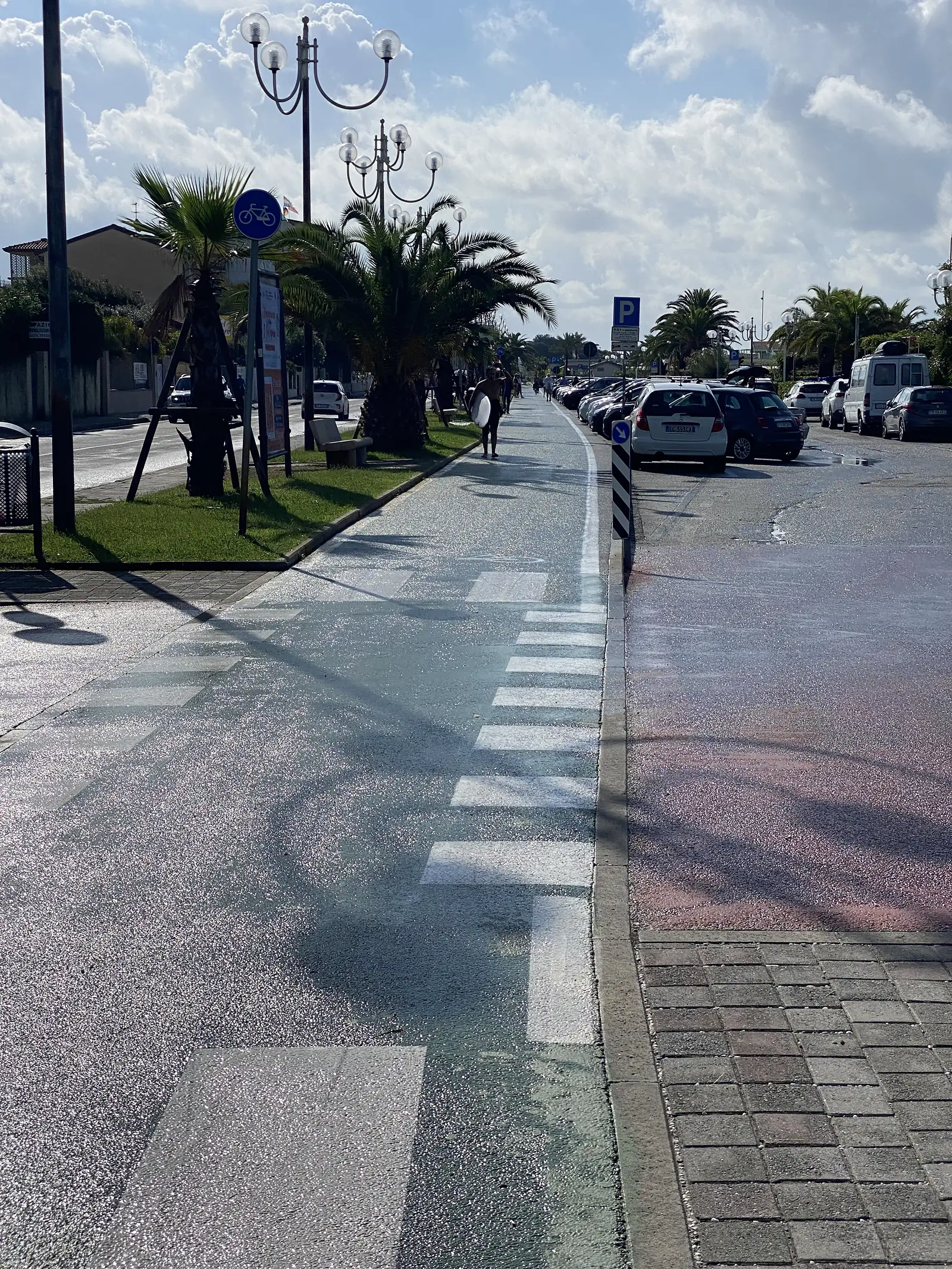 A zebra crossing leads towards a row of ornate lamp posts and palm trees on a paved seafront square, with parked cars and pedestrians nearby.