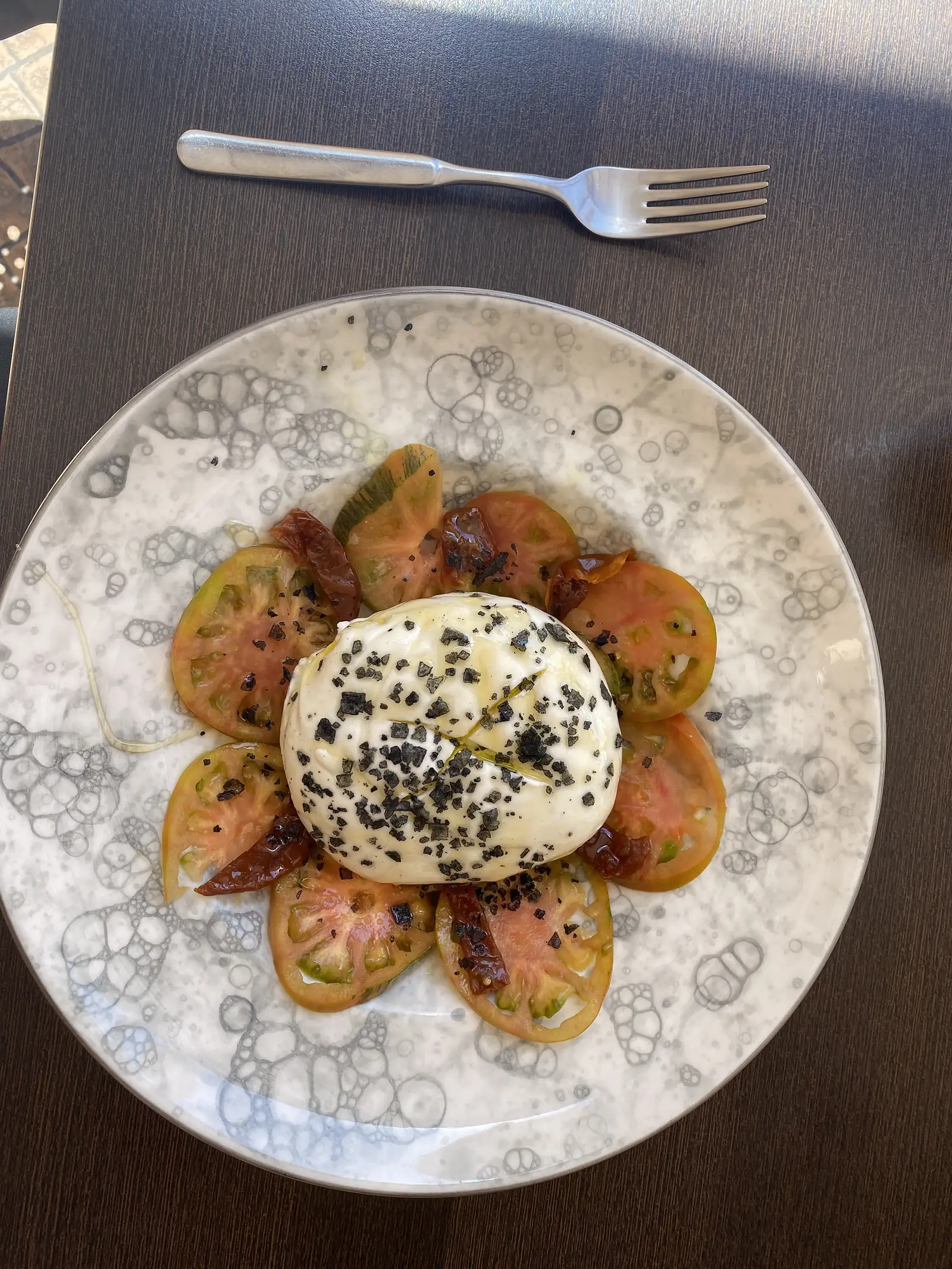 A whole burrata dusted with dark salt flakes on a ring of sliced tomatoes and sun-dried tomatoes on a marbled plate.