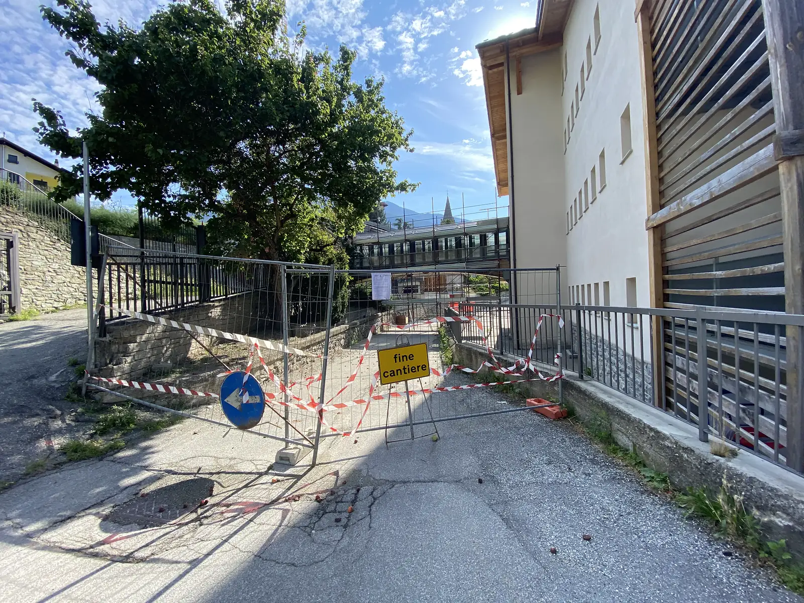 A metal barrier strung with red-and-white tape blocks a path beside a stone building, a yellow sign reading 'fine cantiere' fixed to it.