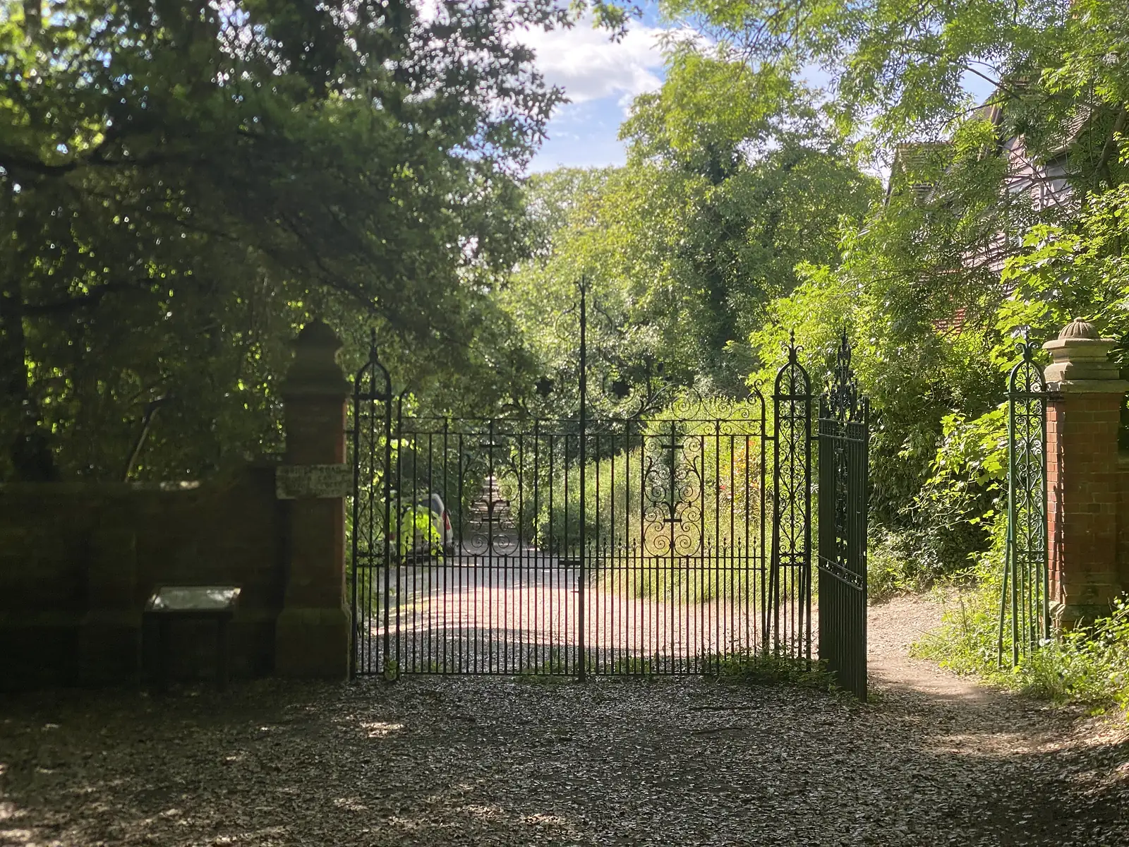 Ornate wrought-iron gates between brick piers standing open onto a shaded gravel drive under big trees.