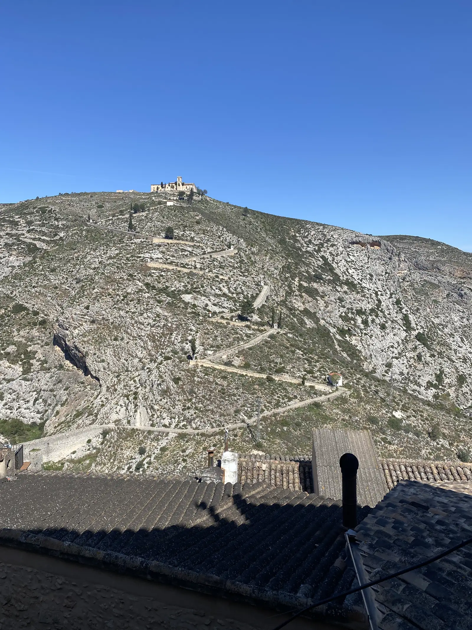 A hilltop sanctuary at the end of a bare limestone ridge with a zigzag walled path climbing to it, seen over the town's rooftops.