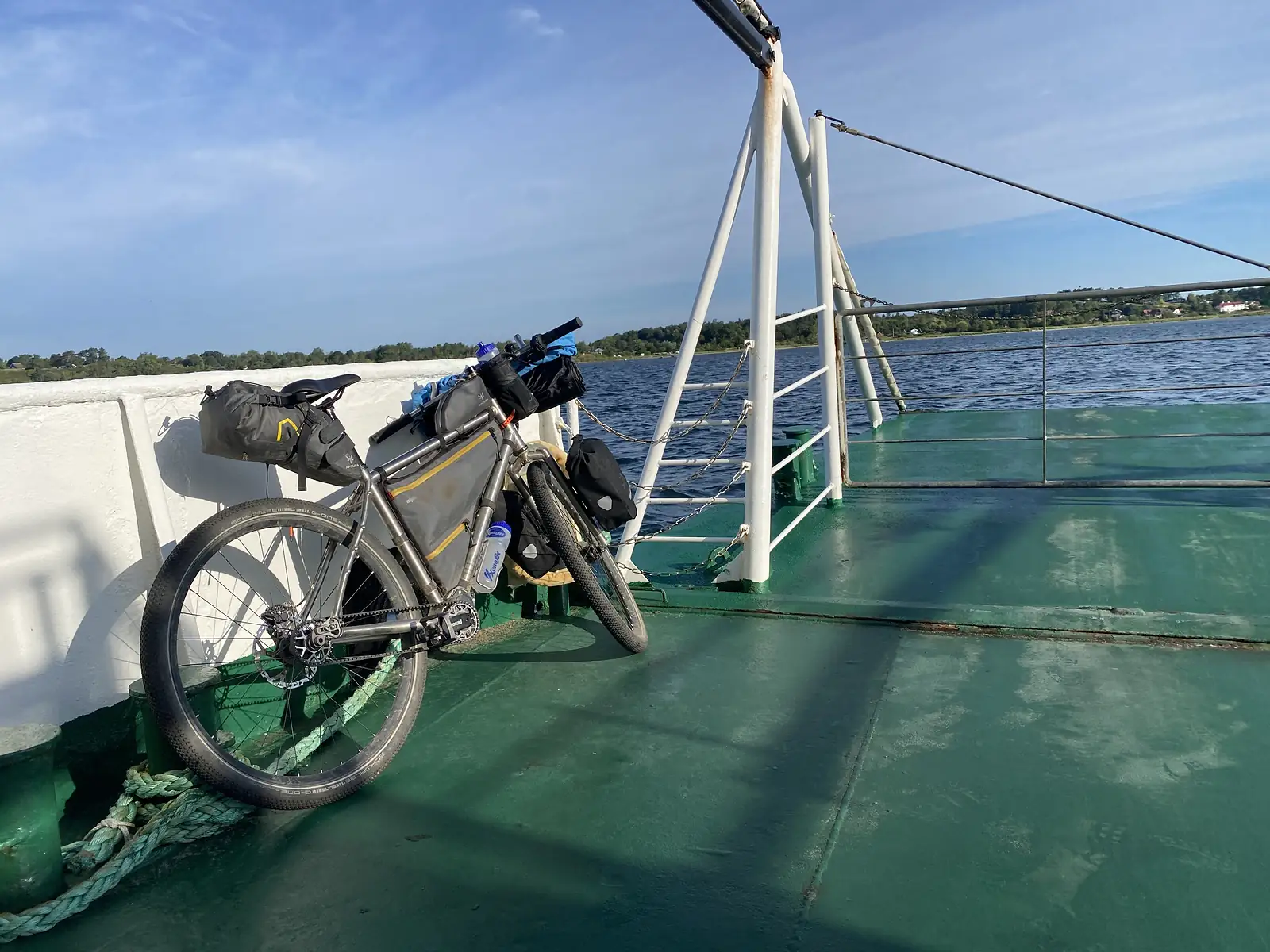 A loaded gravel bike against the rail of a ferry's green deck, a road bridge spanning the water behind.