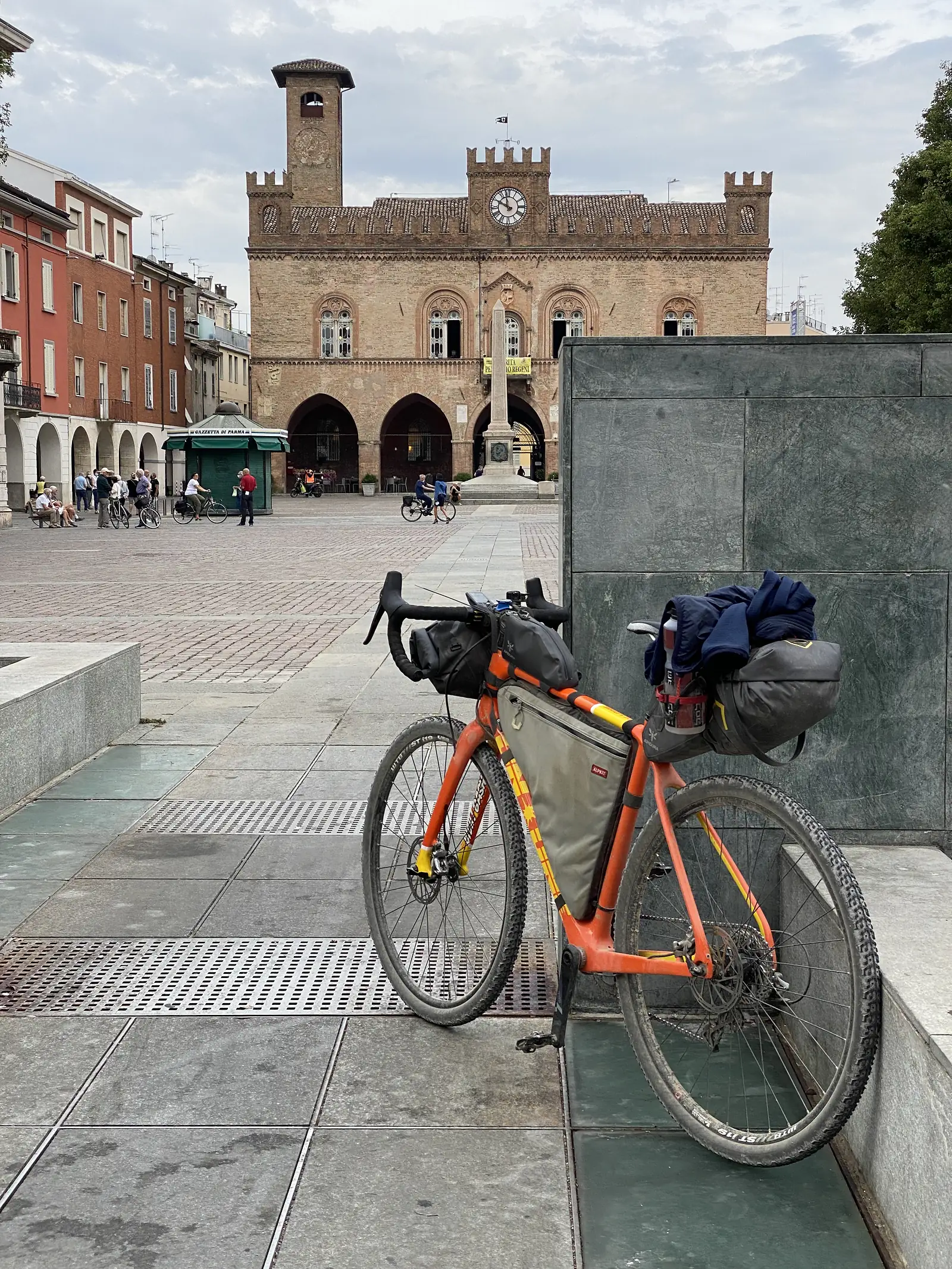 A loaded orange gravel bike parked in a cobbled square below the brick clock tower and cathedral facade of Fidenza.