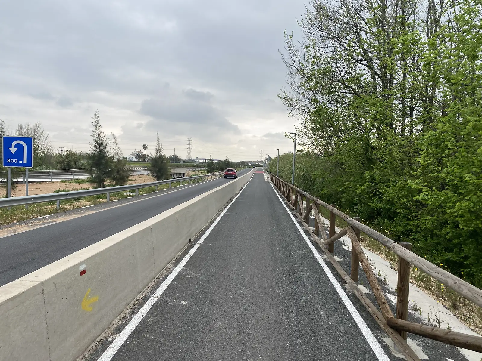 A cycle path divided from the road by a concrete barrier painted with a yellow pilgrim arrow and a red-and-white waymark.